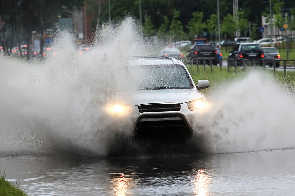 A white SUV driving through a flooded city street with water spraying