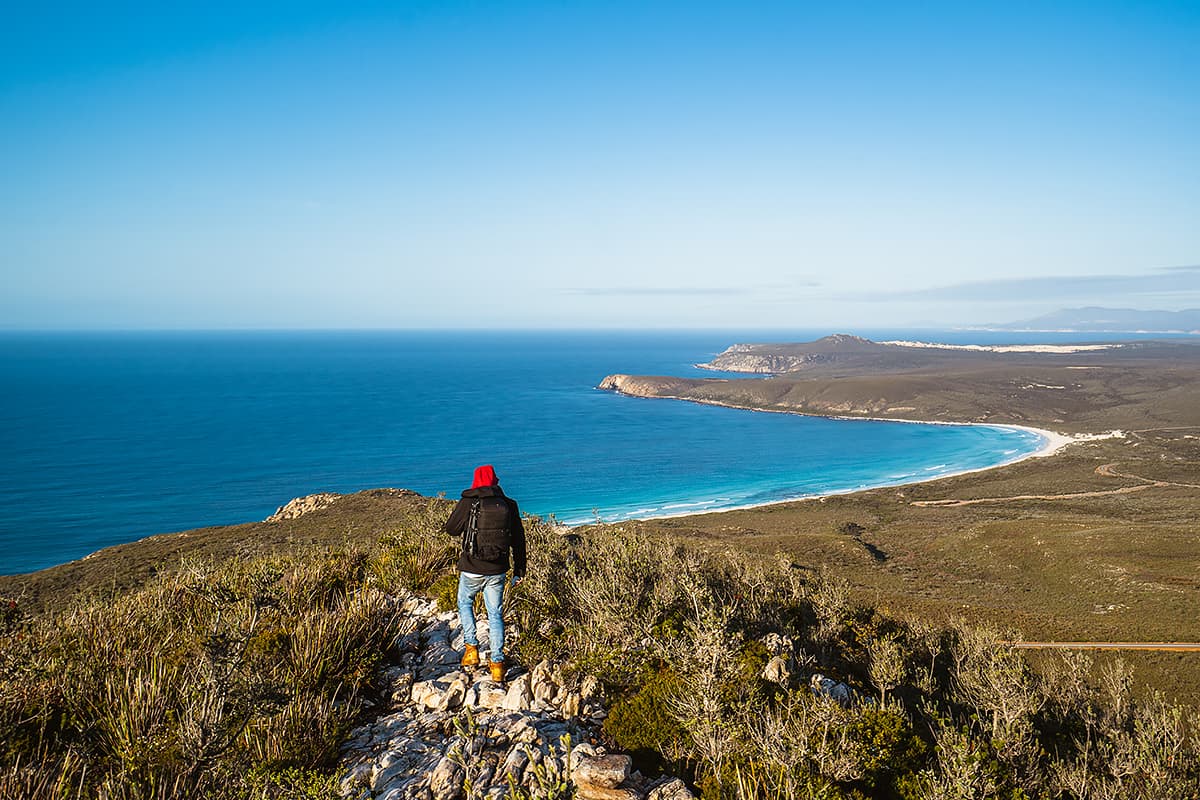 A person on a hiking trail standing on a high peak overlooking the ocean.