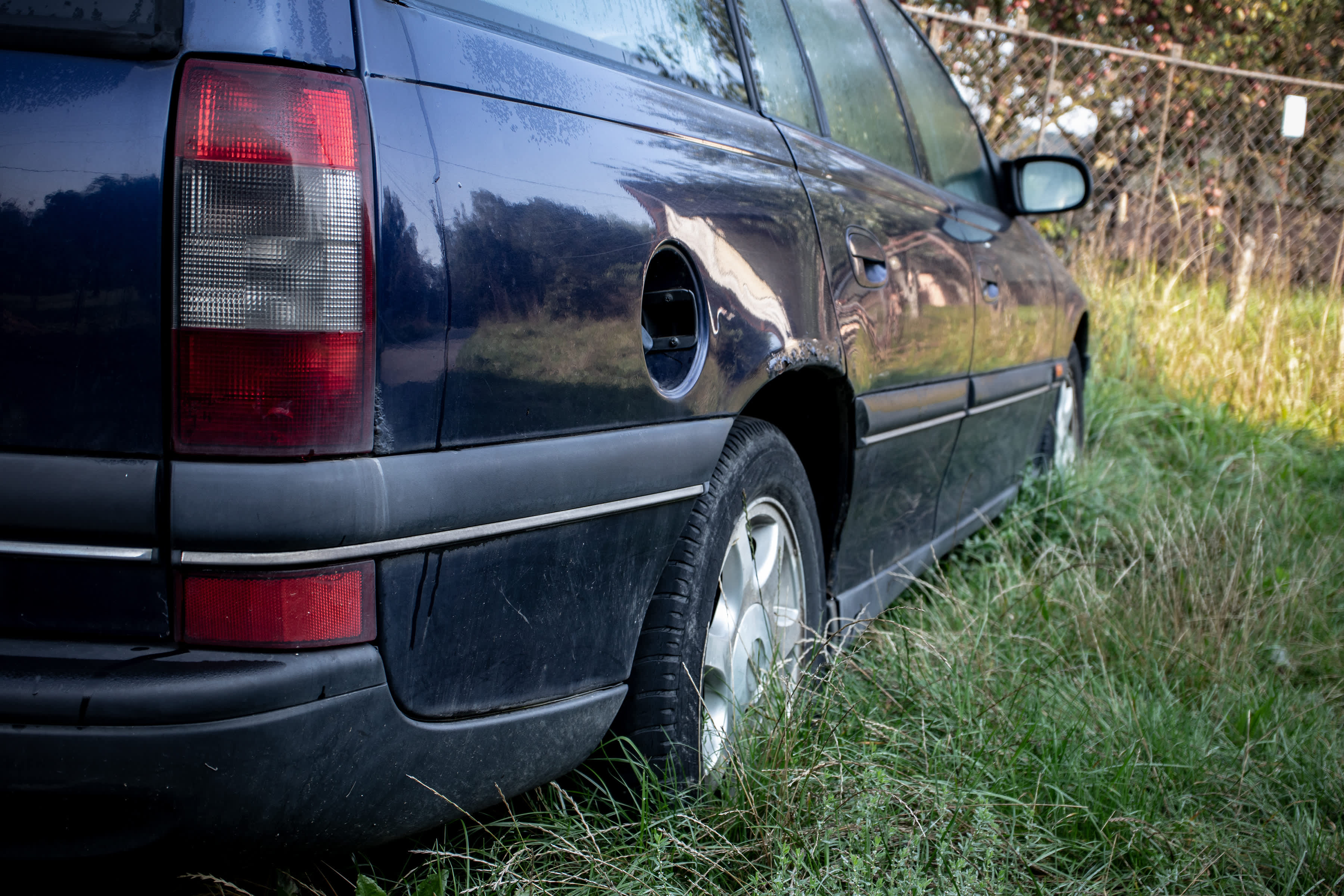 A dark blue station wagon in tall grass near a fence with a missing fuel cap