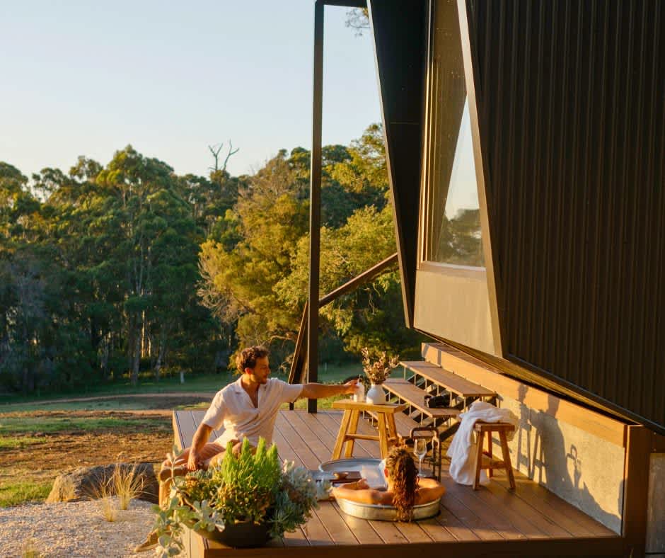 Couple sitting on sunlit deck in front of tiny cabin reading in bathtub and grazing