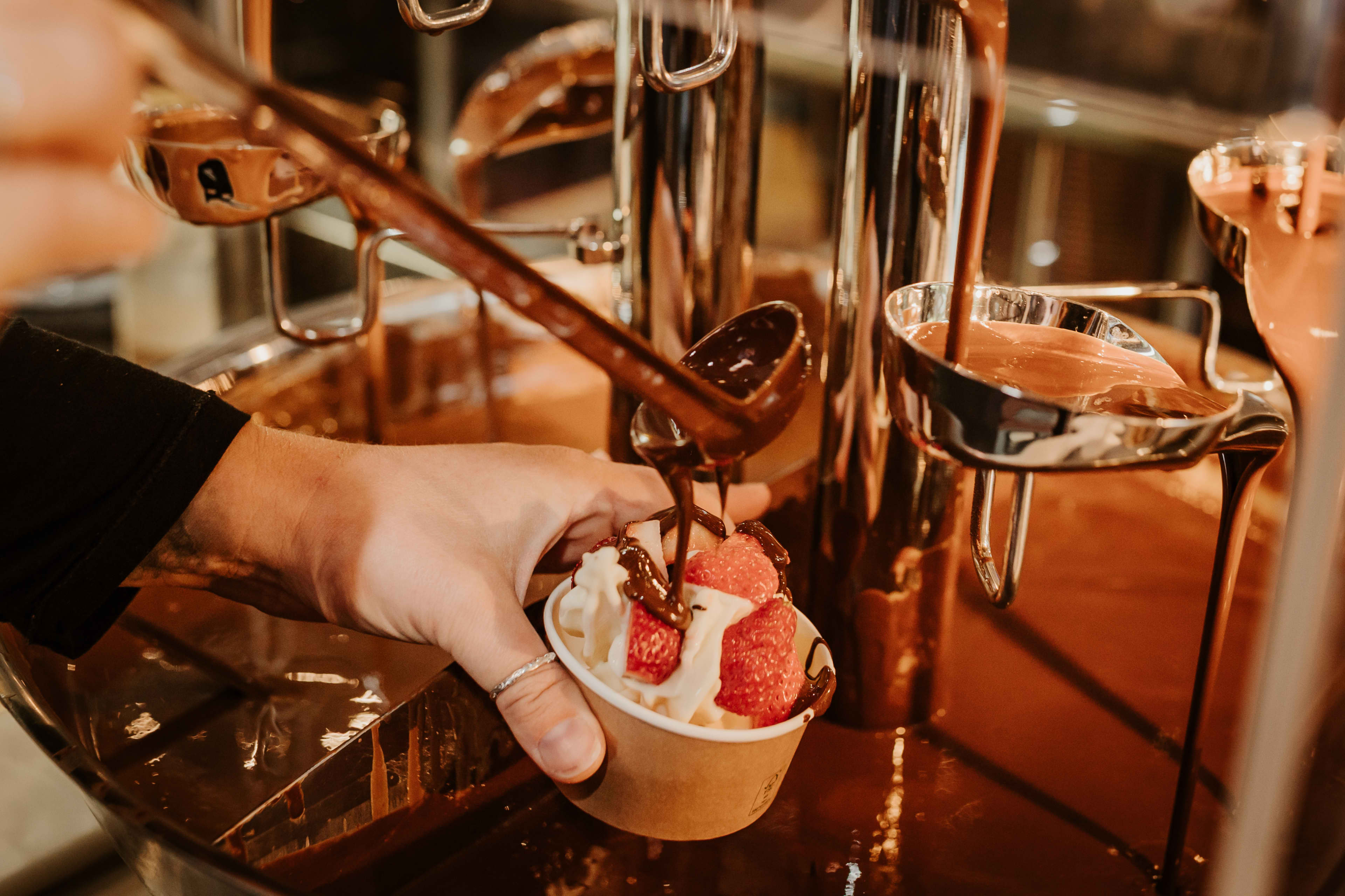 A ladle pouring melted chocolate on a chocolate fountain on to a cup of soft serve ice cream with 3 strawberries.