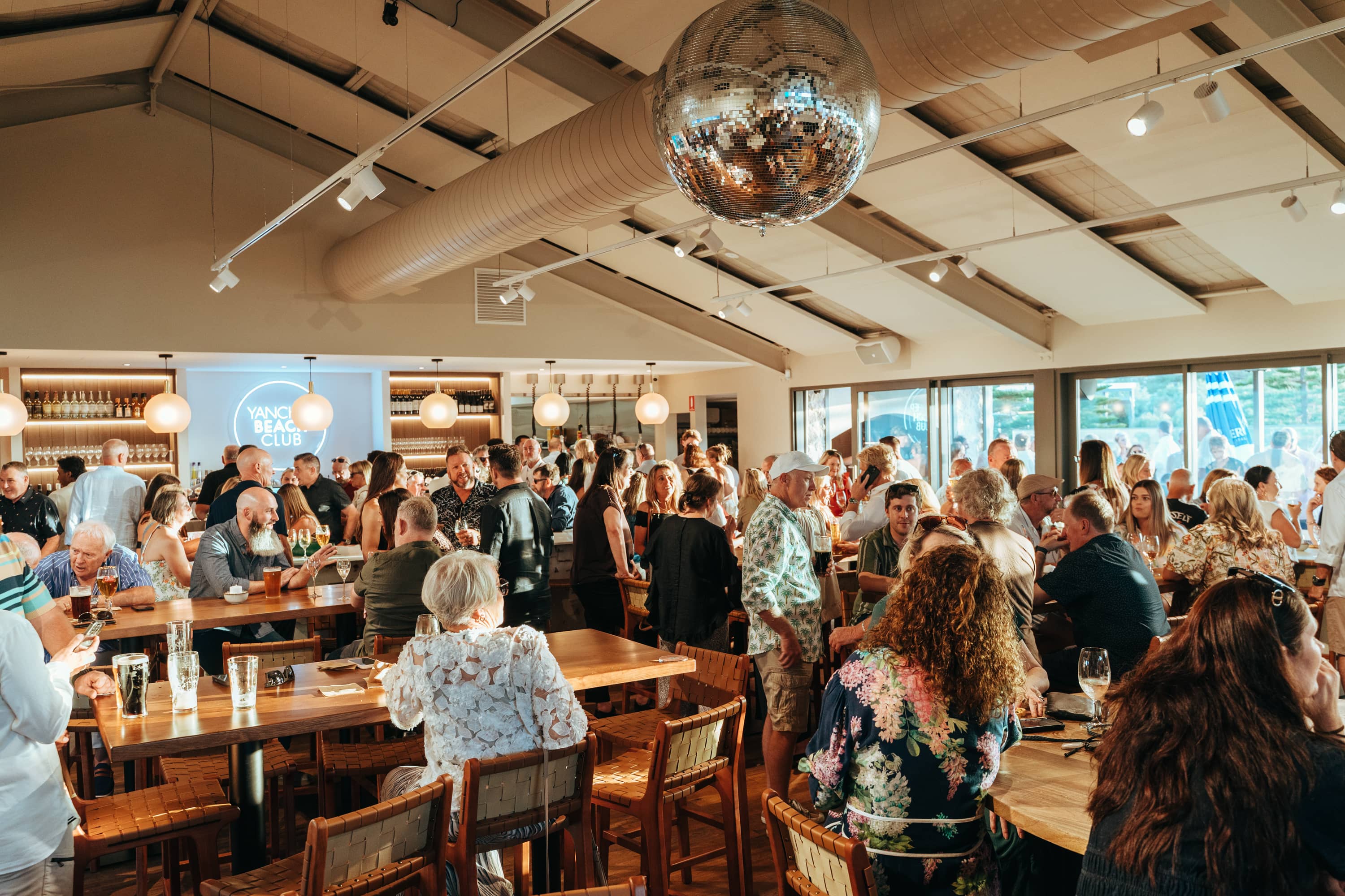 “A crowded, sunlit bar and dining space filled with people socializing around wooden tables and a central bar. Guests of various ages stand and sit holding drinks, chatting in a lively atmosphere. A large mirrored disco ball hangs from the ceiling, with warm pendant lights and exposed beams above.