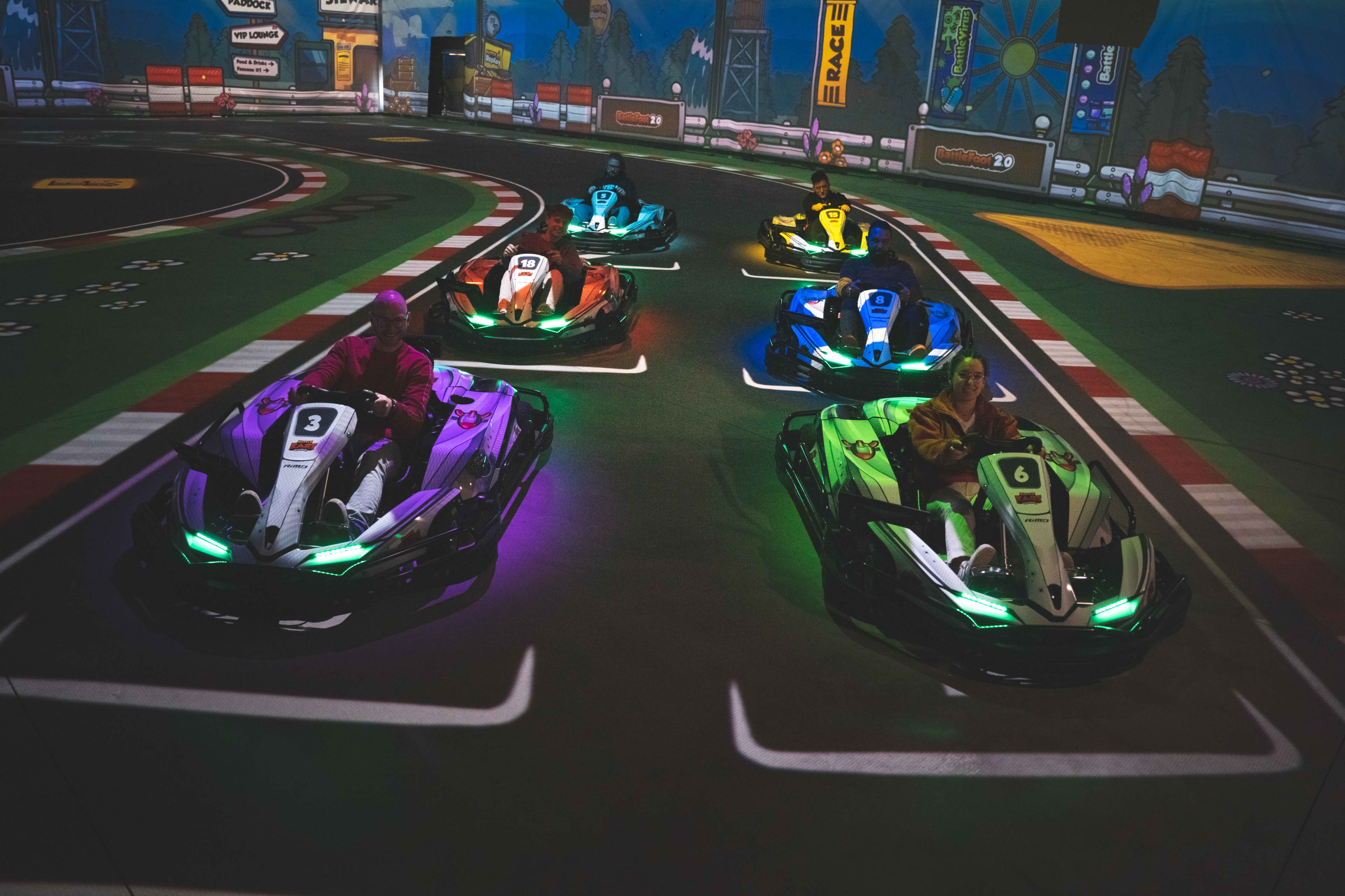 Six people sit in colorful electric go-karts lined up on an indoor racetrack, viewed from the front. The karts have glowing LED lights and visible numbers, positioned on starting grid lines with a curving track, barriers, and race-themed decorations in the background.
