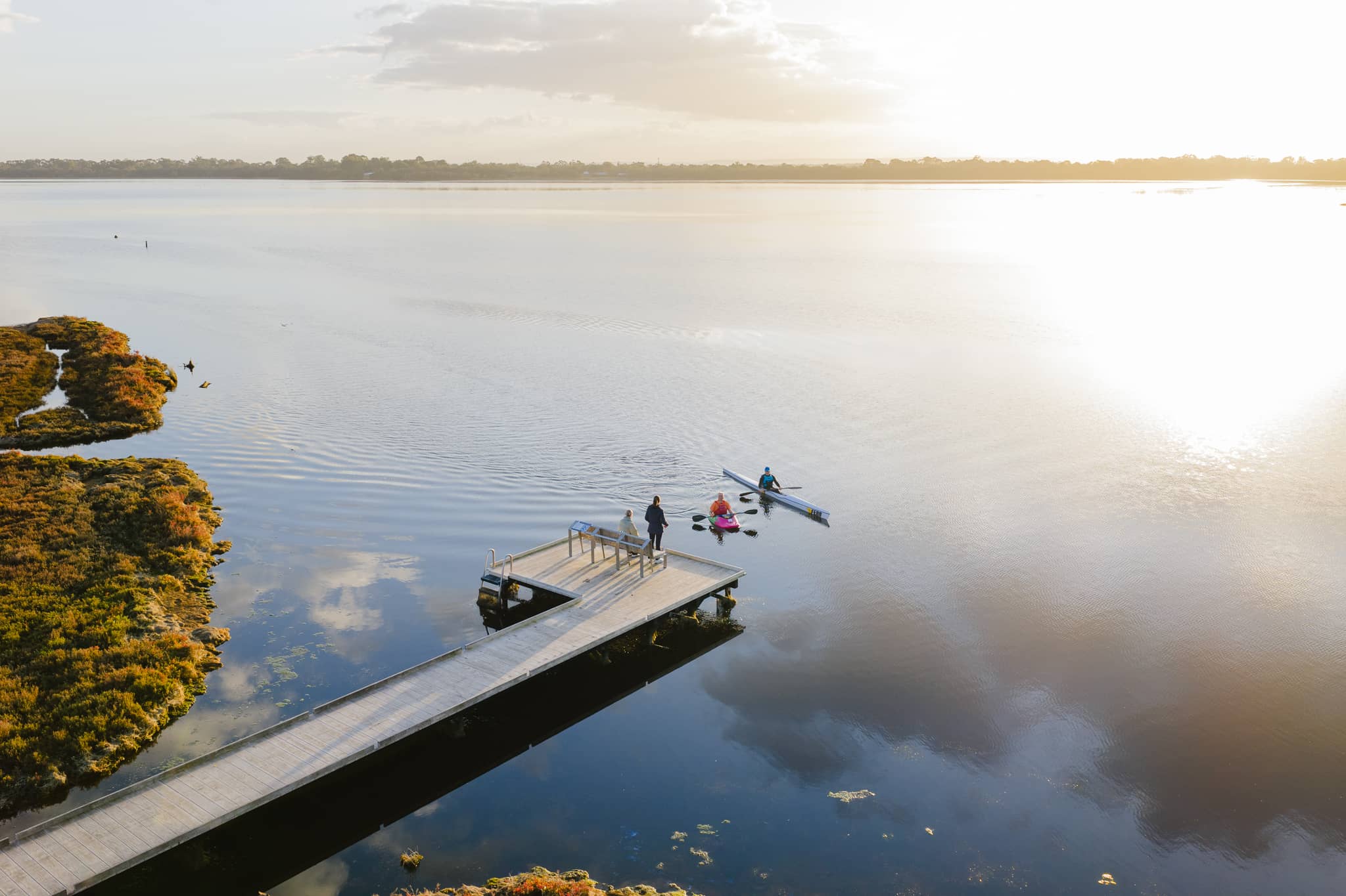 An aerial view of two people canoeing at sunrise near a wooden boardwalk, while two others stand on the boardwalk overlooking a calm estuary.