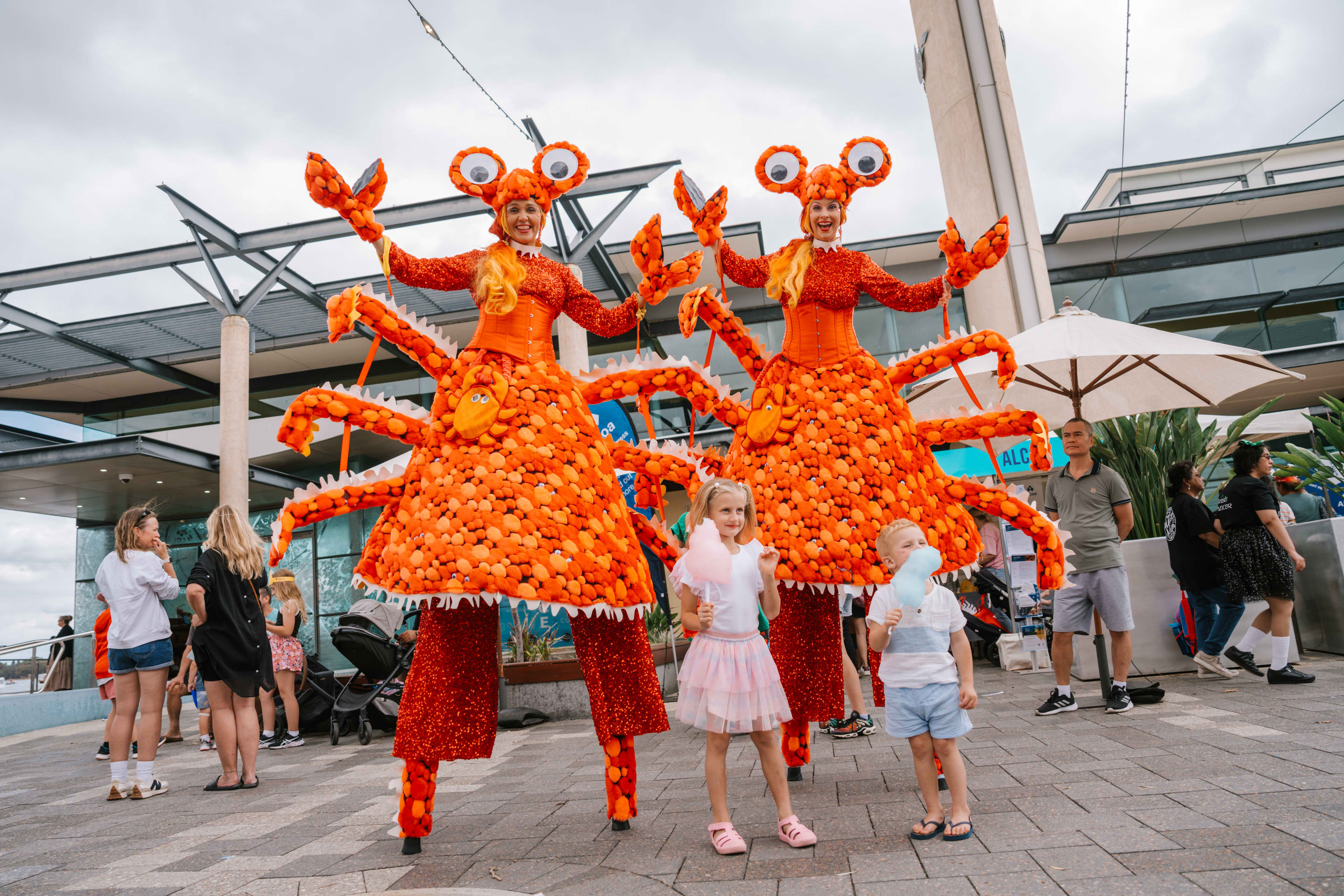 Two performers dressed in bright orange crab costumes on tall stilts pose with raised claws at an outdoor waterfront event, while two young children in front hold cotton candy and look toward the camera, with a small crowd and modern buildings in the background.