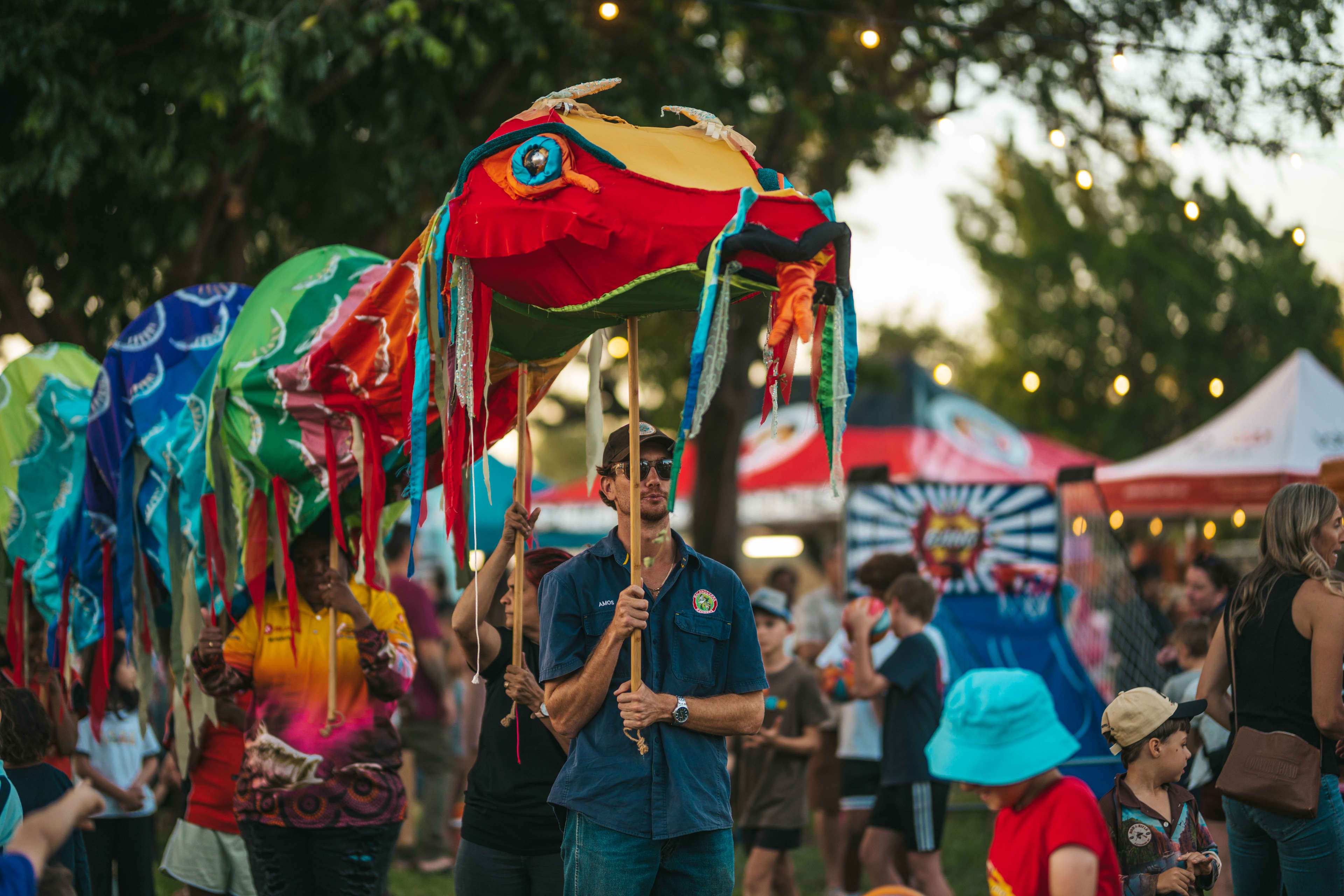 A man carrying a large, colourful puppet decorated with ribbons through a lively festival crowd.
