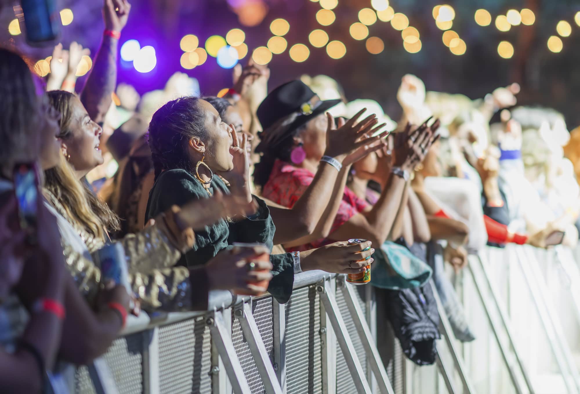 A crowd cheering at the fenceline of a stage, with lights glowing in the distance.