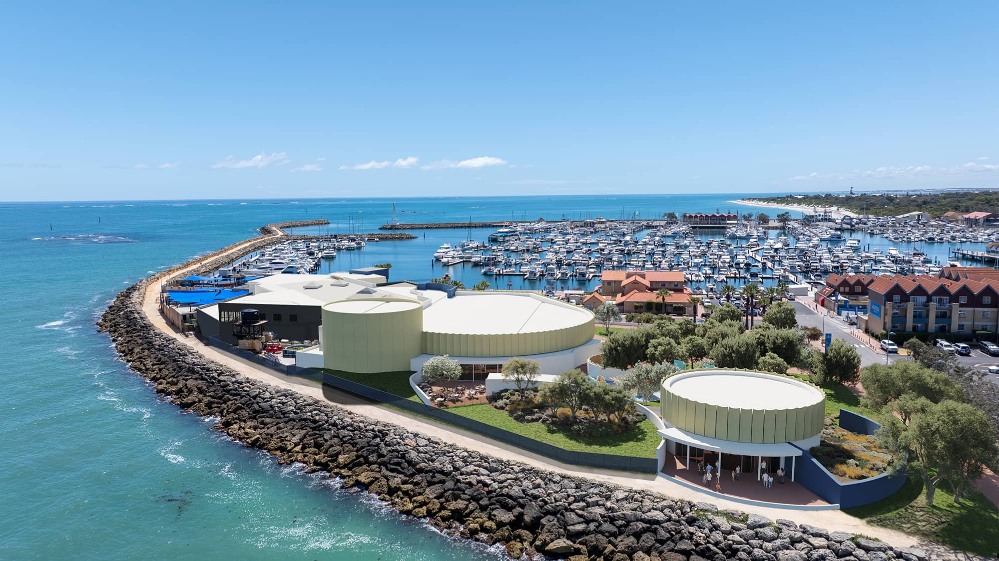 Aerial view of a coastal marina with turquoise water and numerous docked boats. In the foreground, a modern waterfront complex with curved, light-colored cylindrical buildings sits on a rocky breakwater surrounded by landscaped greenery and walking paths. The marina extends into the distance with rows of white yachts, red-roofed buildings along the shore, and a sandy coastline under a clear blue sky.
