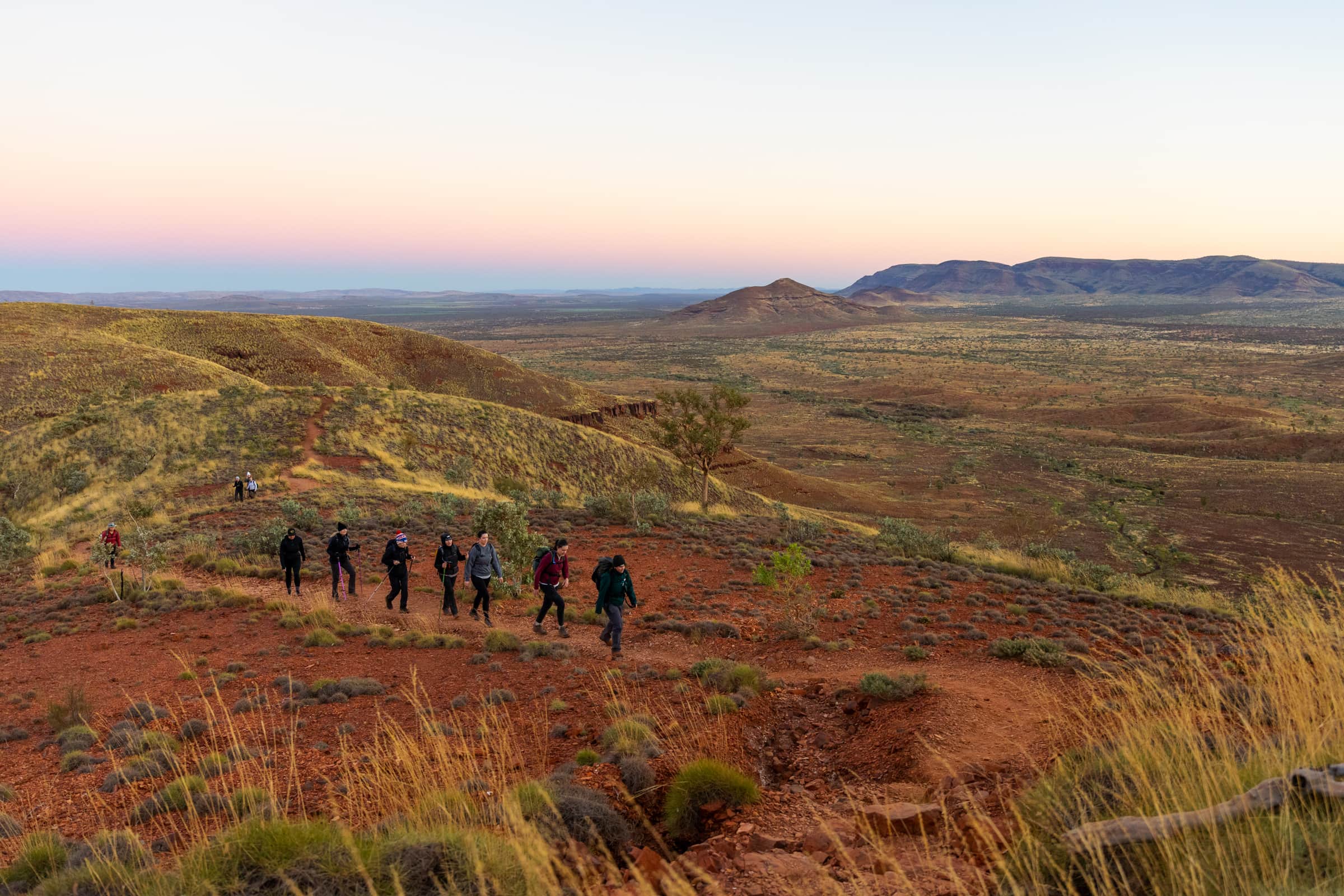 A group of hikers walking up a rocky terrain complete with spinifex, and rugged, red cliffs towering in the background.