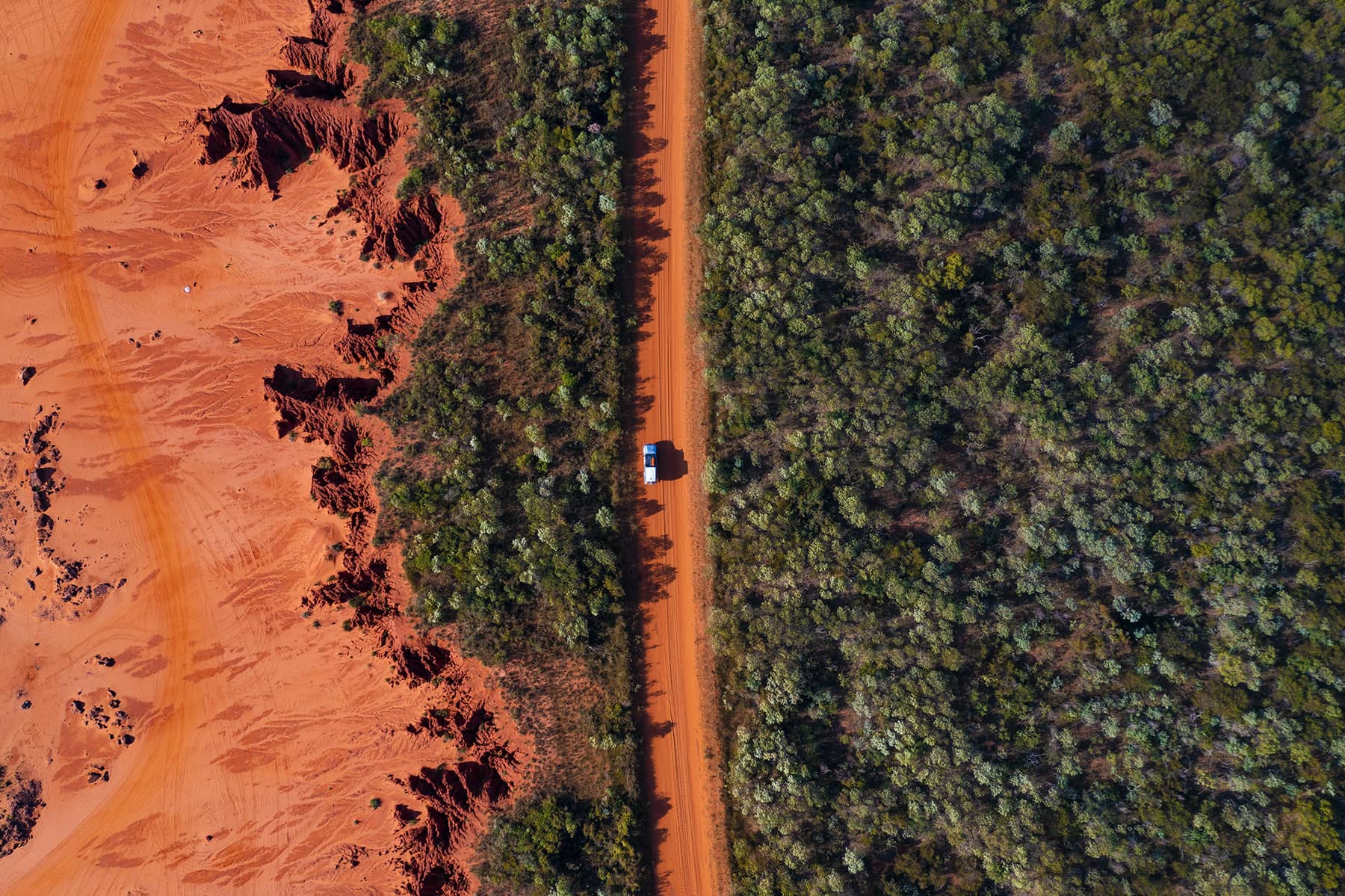 A red desert plain with cliff edges with a car driving down an unsealed track surrounded by green shrubs.