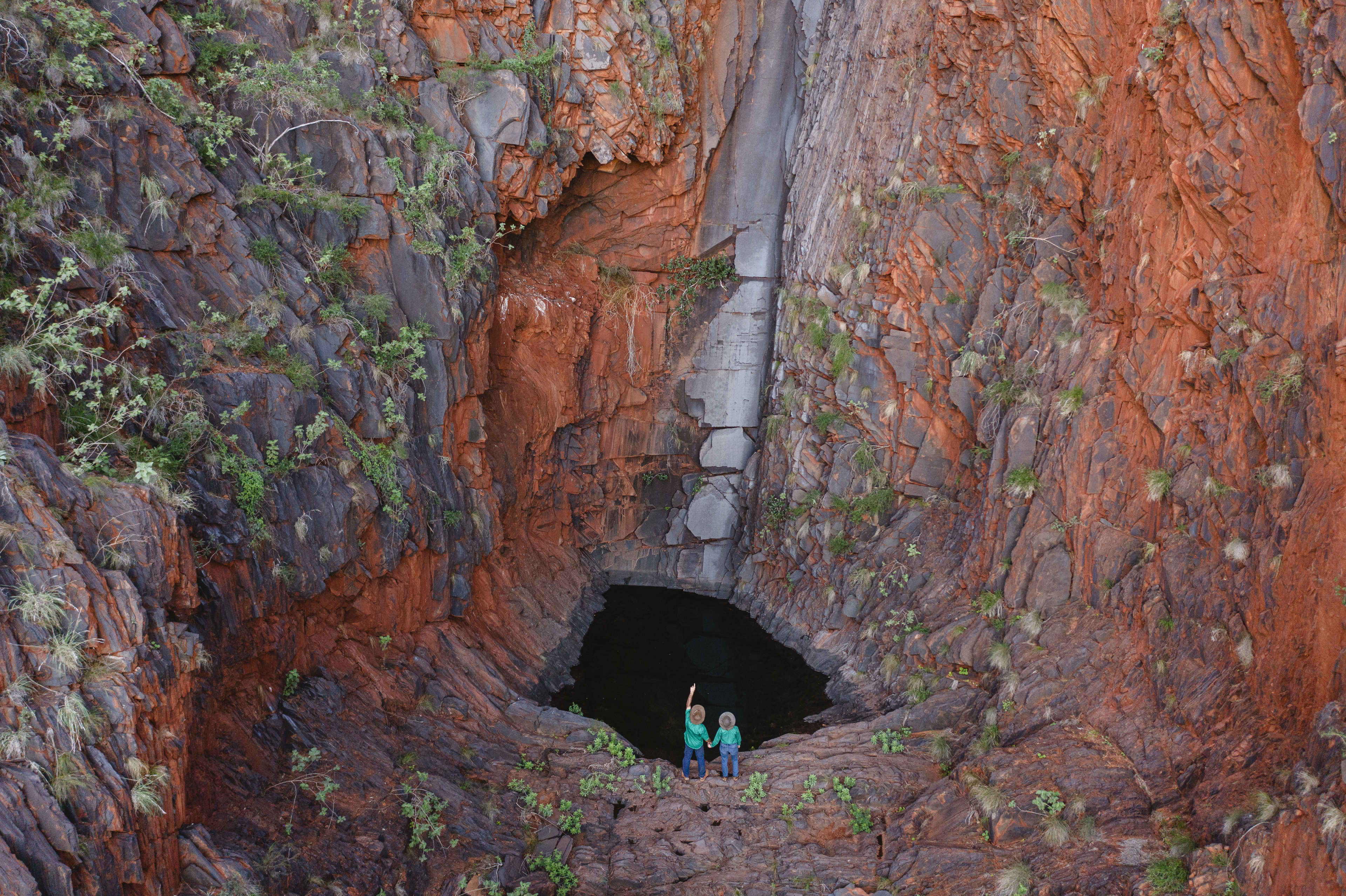 An aerial image of a couple wearing green station shirts and a wide brimmed hat, standing in a deep cut gorge with a small watering hole.