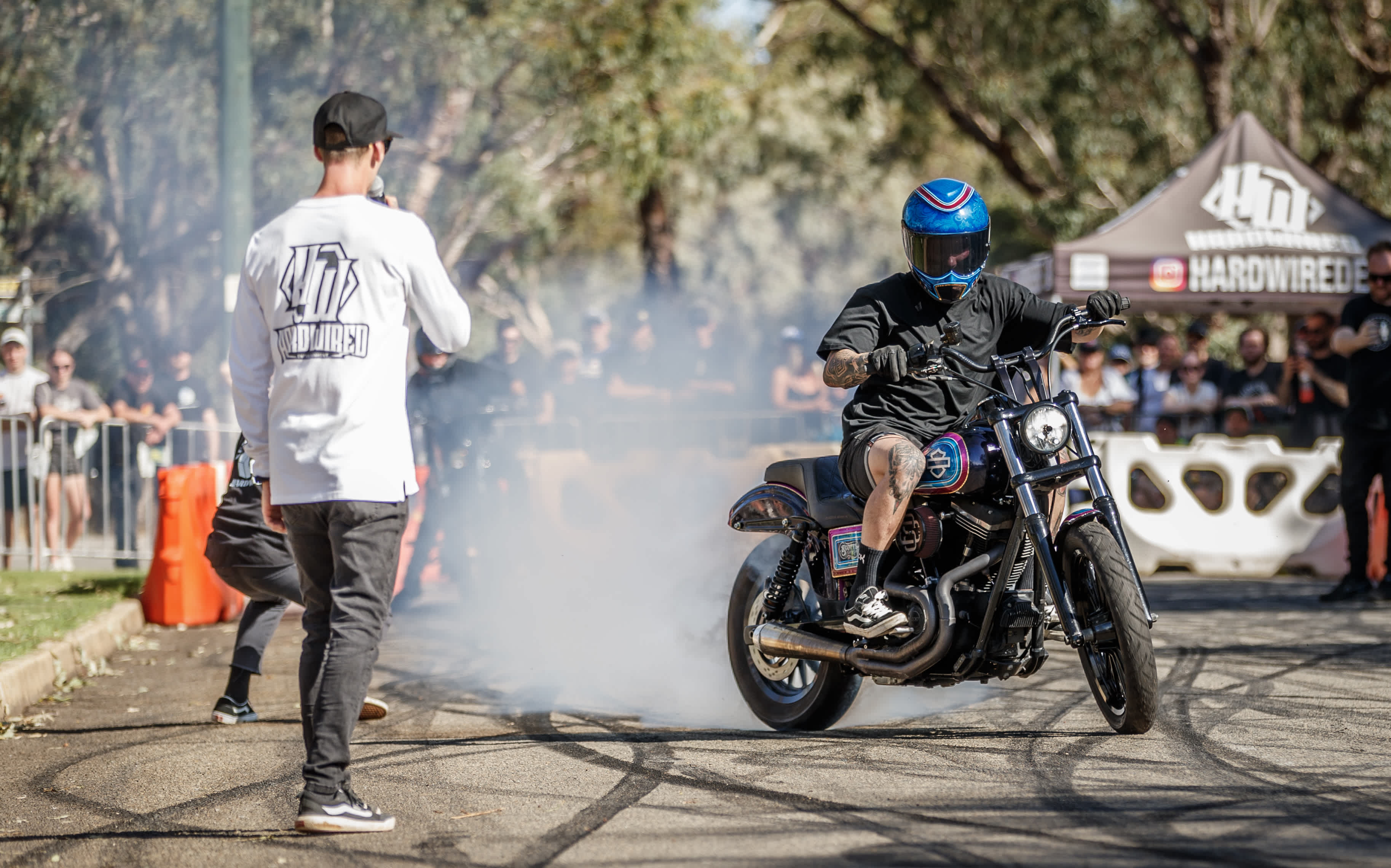 A motorcyclist wearing a blue helmet performs a burnout on a custom motorcycle, creating smoke on a paved area while an announcer with a microphone stands nearby and spectators watch behind barriers at an outdoor event.