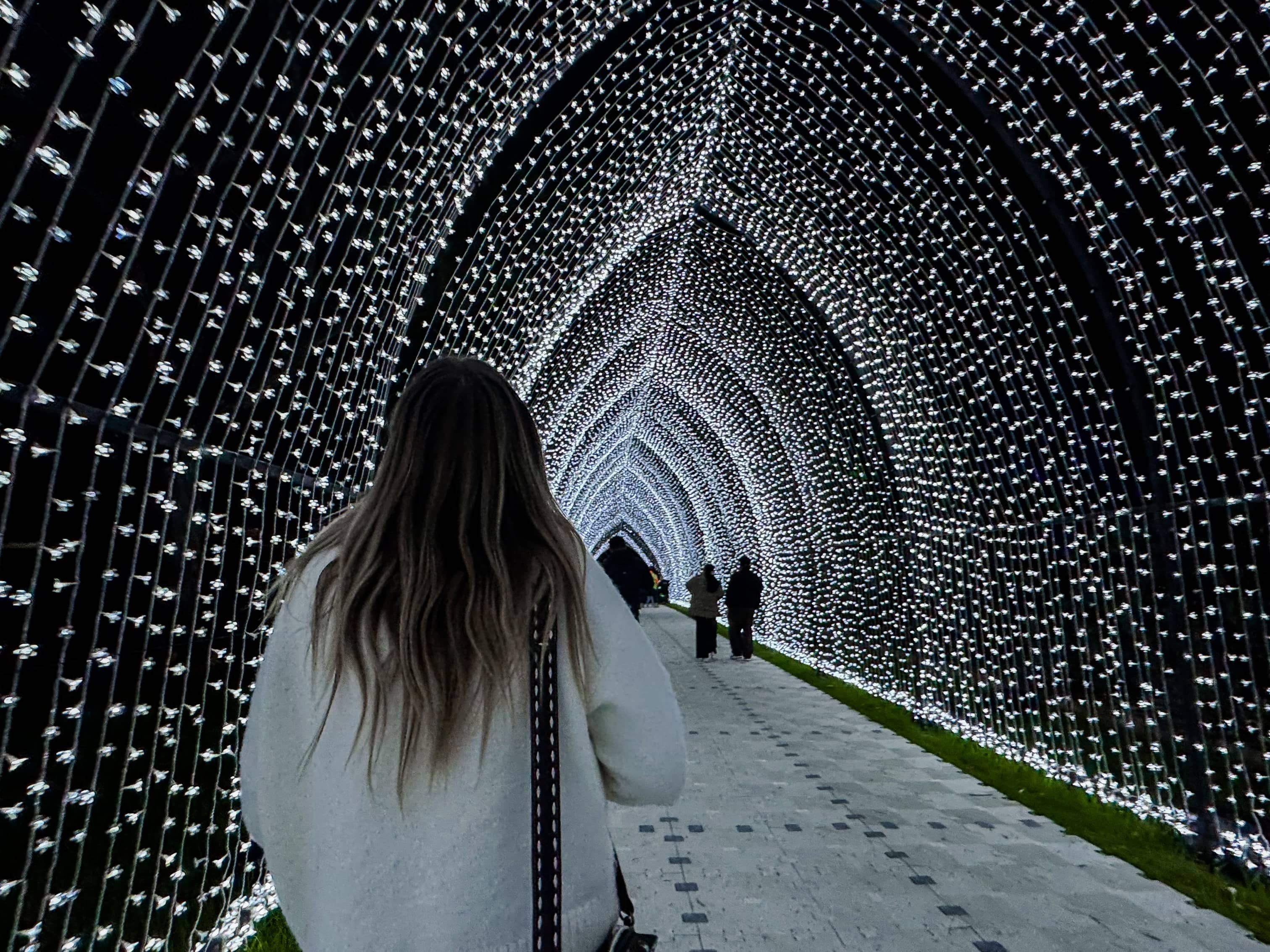 Person walking through an arched tunnel of white lights at night, with illuminated strands forming a glowing canopy overhead and other visitors visible in the distance.