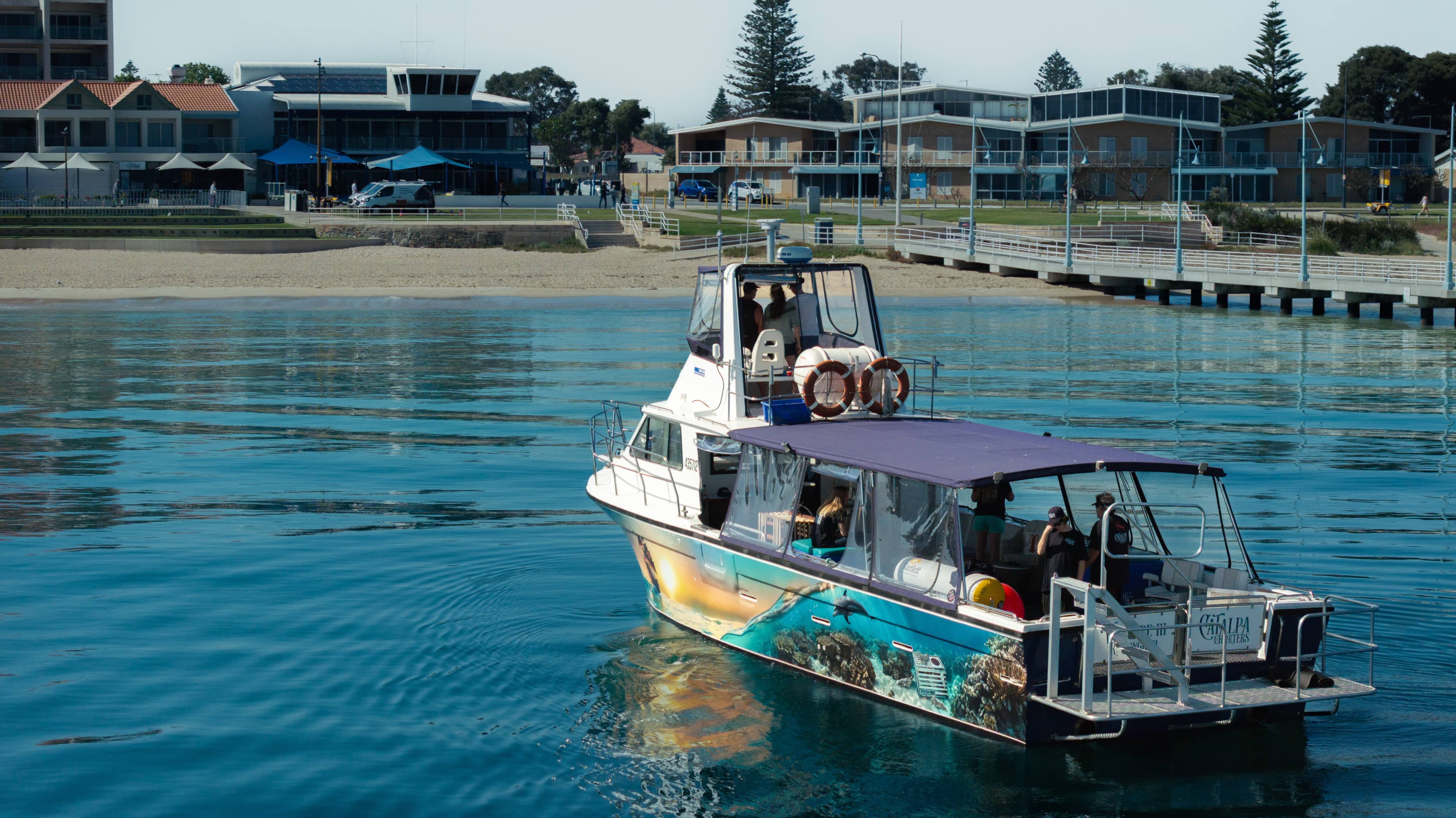 A small tour boat with a colorful ocean-themed mural on its side moves through calm blue water near a waterfront. Several people are on board under a canopy, and life rings are mounted on the upper deck. In the background, there are modern low-rise buildings, a pier with railings, and a sandy shoreline lined with trees.