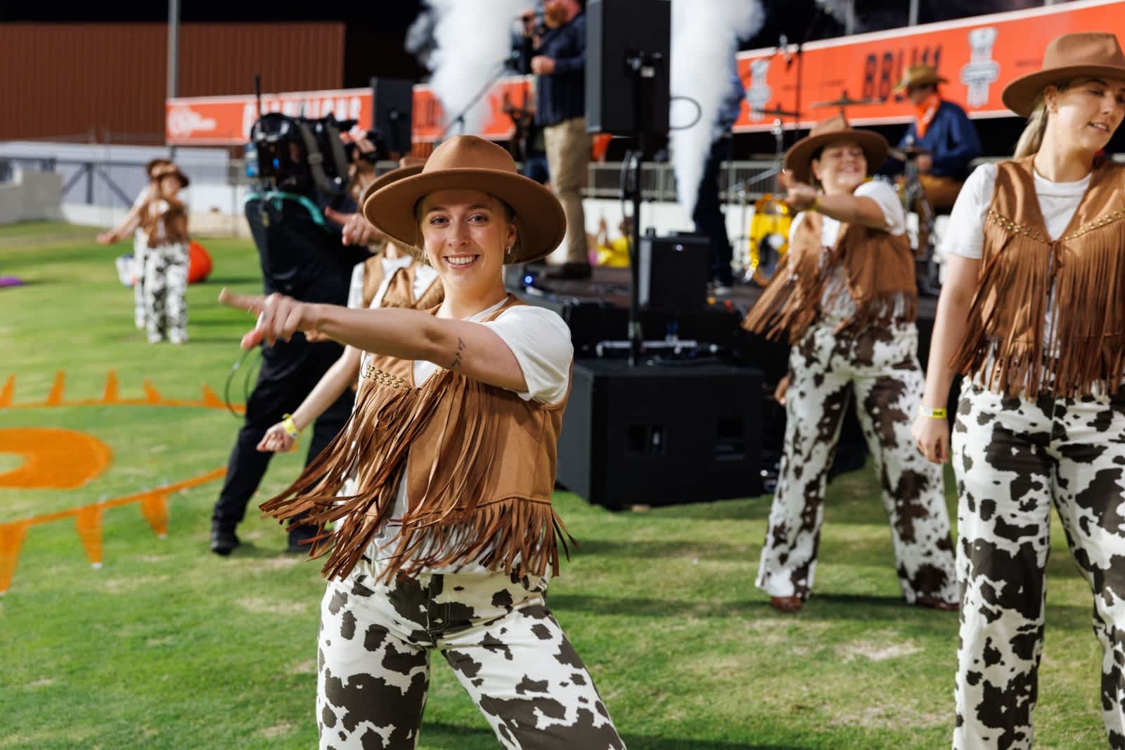 A group of dancers in western-themed outfits perform on a grassy outdoor stage. In the foreground, a smiling woman wearing a brown cowboy hat, fringed vest, white shirt, and cow-print pants points toward the camera while dancing. Behind her, other similarly dressed dancers move in sync, with a live band playing on a stage in the background.