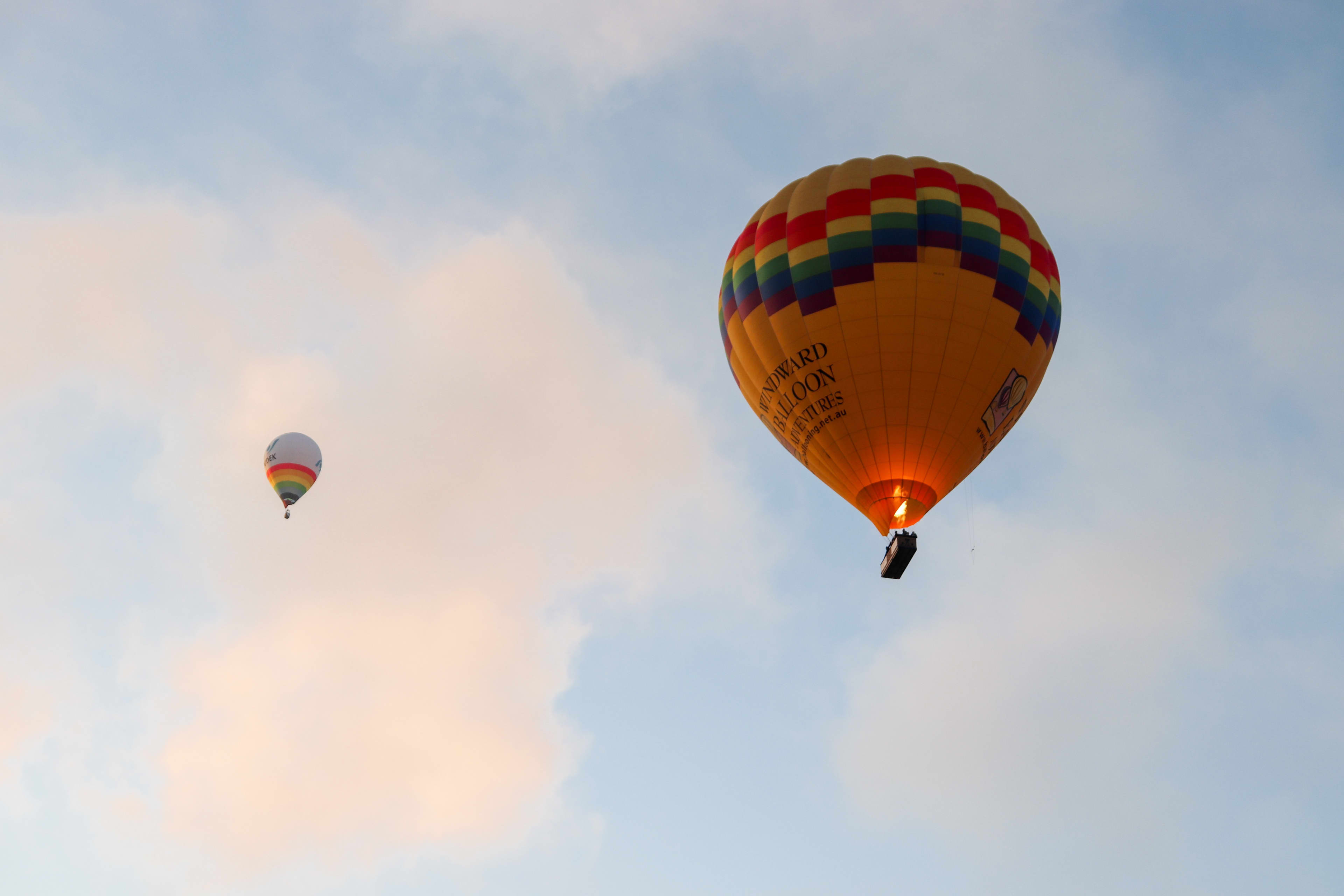 A large, yellow hot air balloon with a rainbow pattern around its top rim floats in a soft blue sky. The balloon's burner glows brightly with an orange flame, illuminating the basket below. Text on the side of the yellow envelope reads "Windward Balloon Adventures". Further away in the distance, a smaller white hot air balloon with a rainbow stripe floats against wispy, pastel pink and white clouds.
