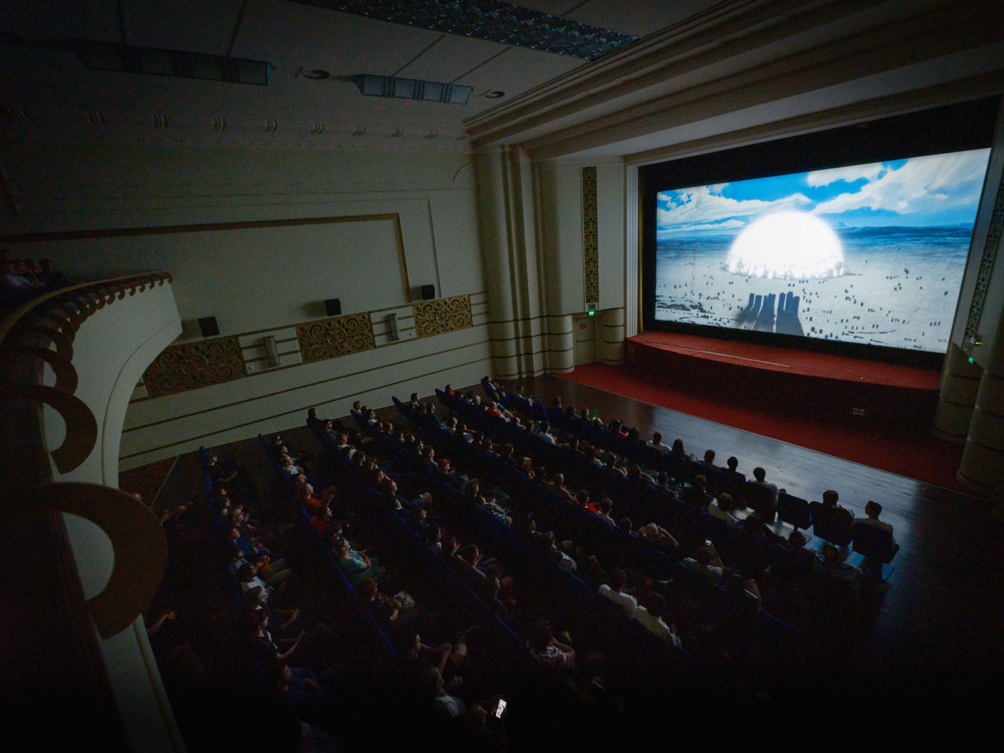A high-angle view from a balcony inside a historic movie theater filled with an audience watching a film. A large, bright screen at the front displays an animated scene featuring a large white dome or sun on a horizon under a blue, cloudy sky. The theater's interior features classic architectural detailing, including a grand proscenium arch framing the screen and decorative panels along the side walls.