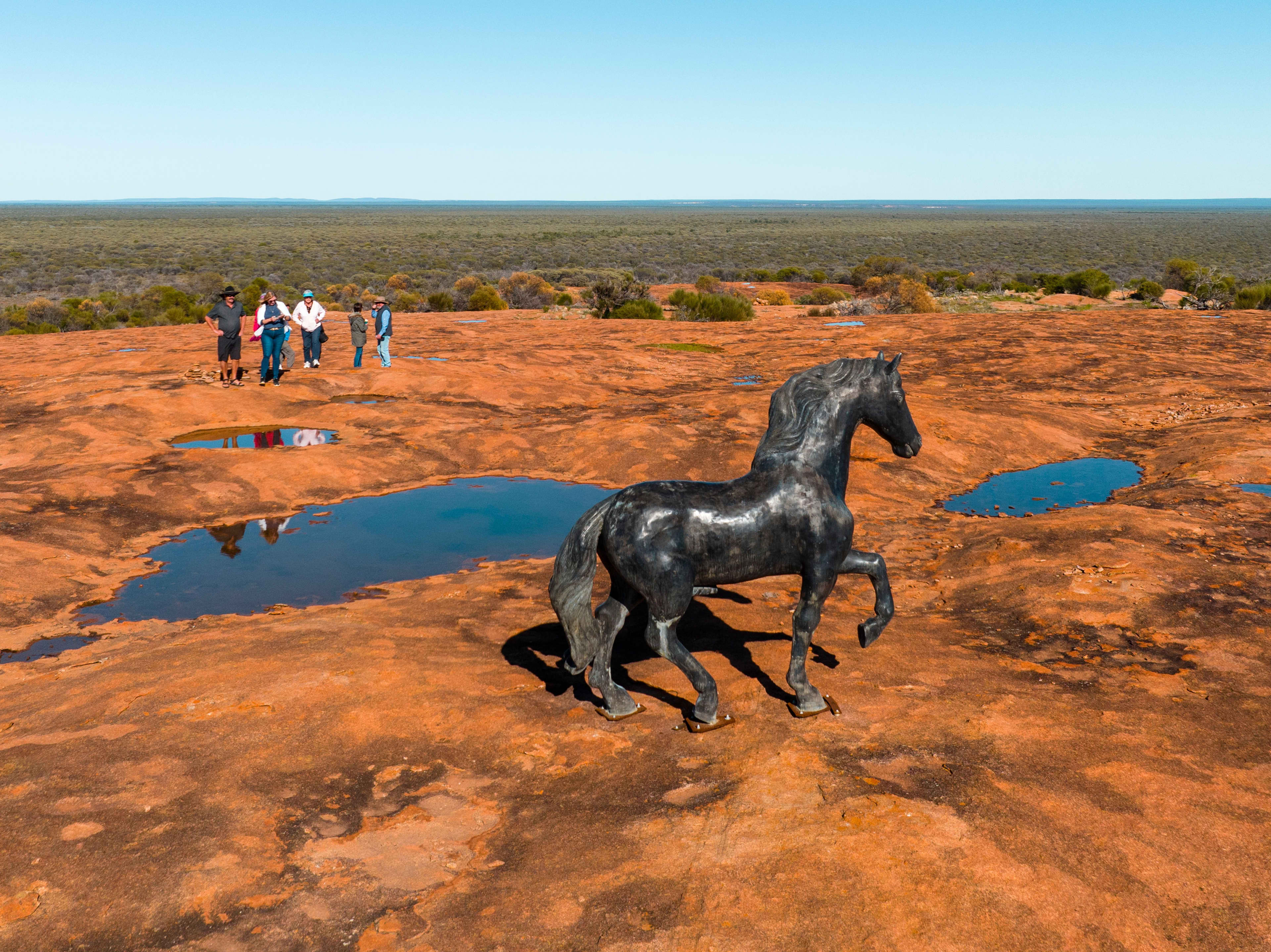 A black horse sculpture stands on a wide orange-red rock plateau dotted with shallow puddles, while a small group of people walks in the background under a clear blue sky. Sparse scrubland stretches to the horizon, giving the scene a remote desert-like atmosphere.