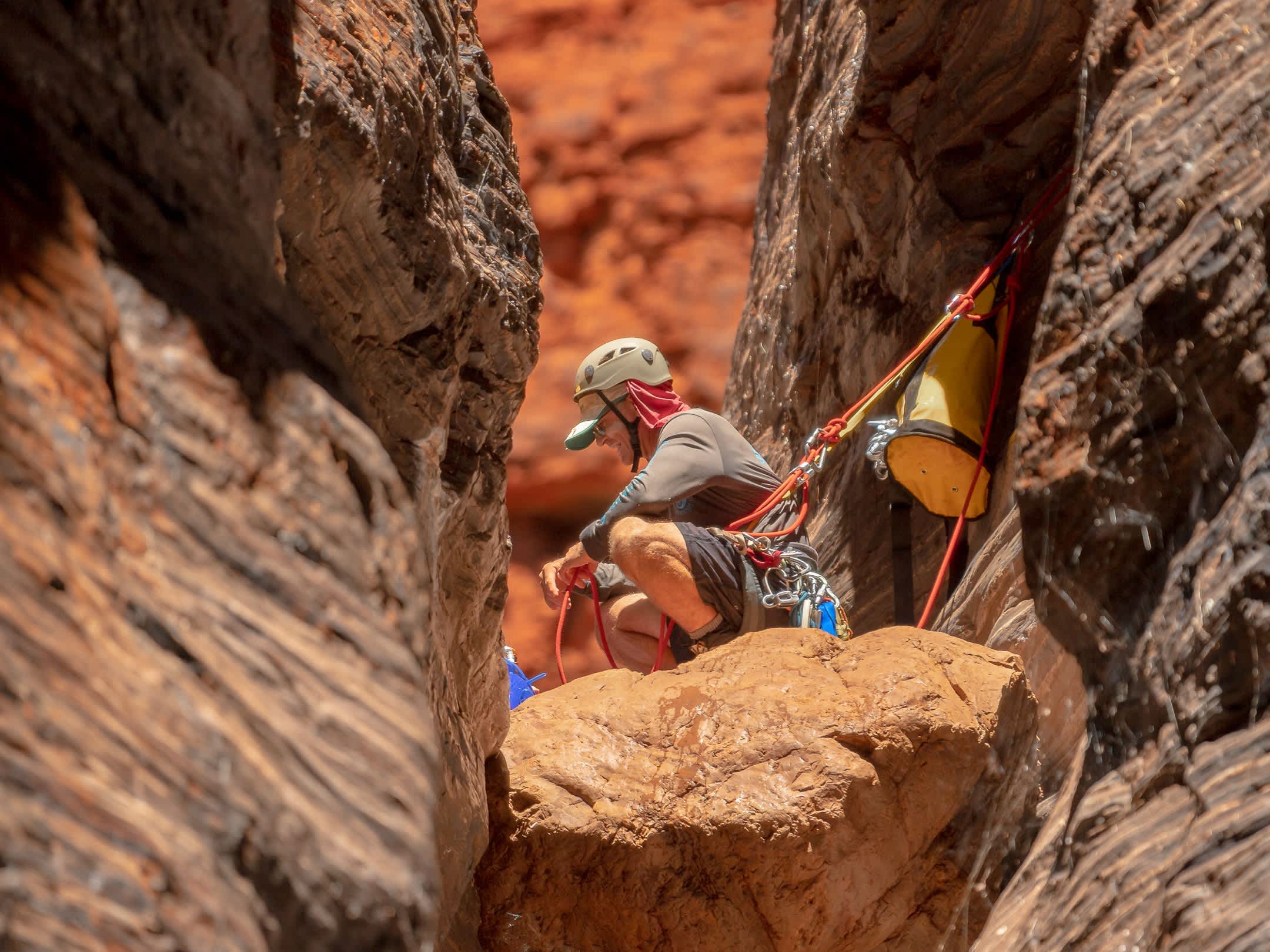 A close-up view of a canyoneer wearing a helmet and safety harness crouching on a large boulder wedged securely within a tight, sun-drenched red rock canyon crevice.