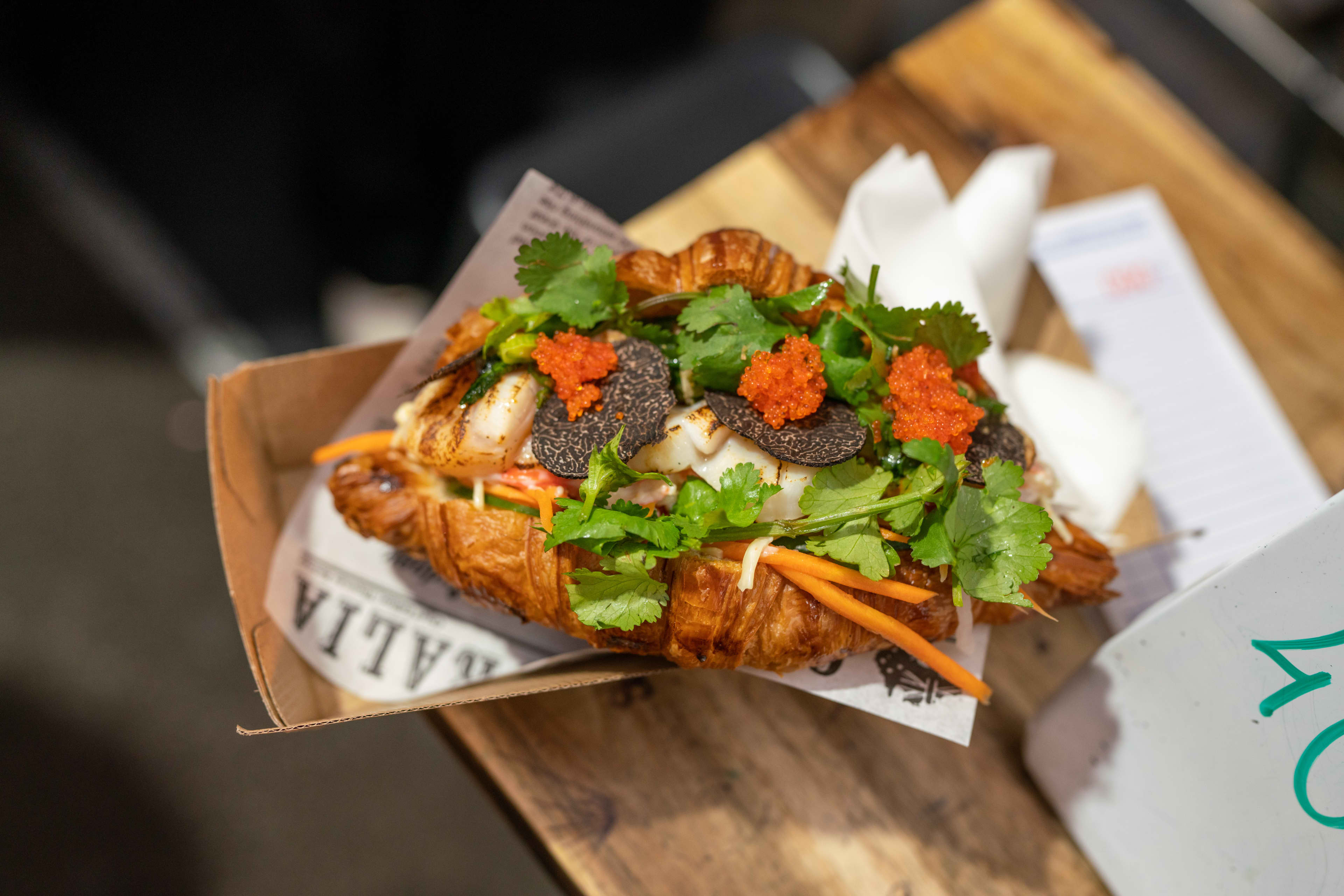 A savory croissant sandwich sits in a brown cardboard tray on a wooden surface, overflowing with fillings including fresh cilantro sprigs, shredded carrots, bright orange tobiko fish roe, and thin slices of black truffle.
