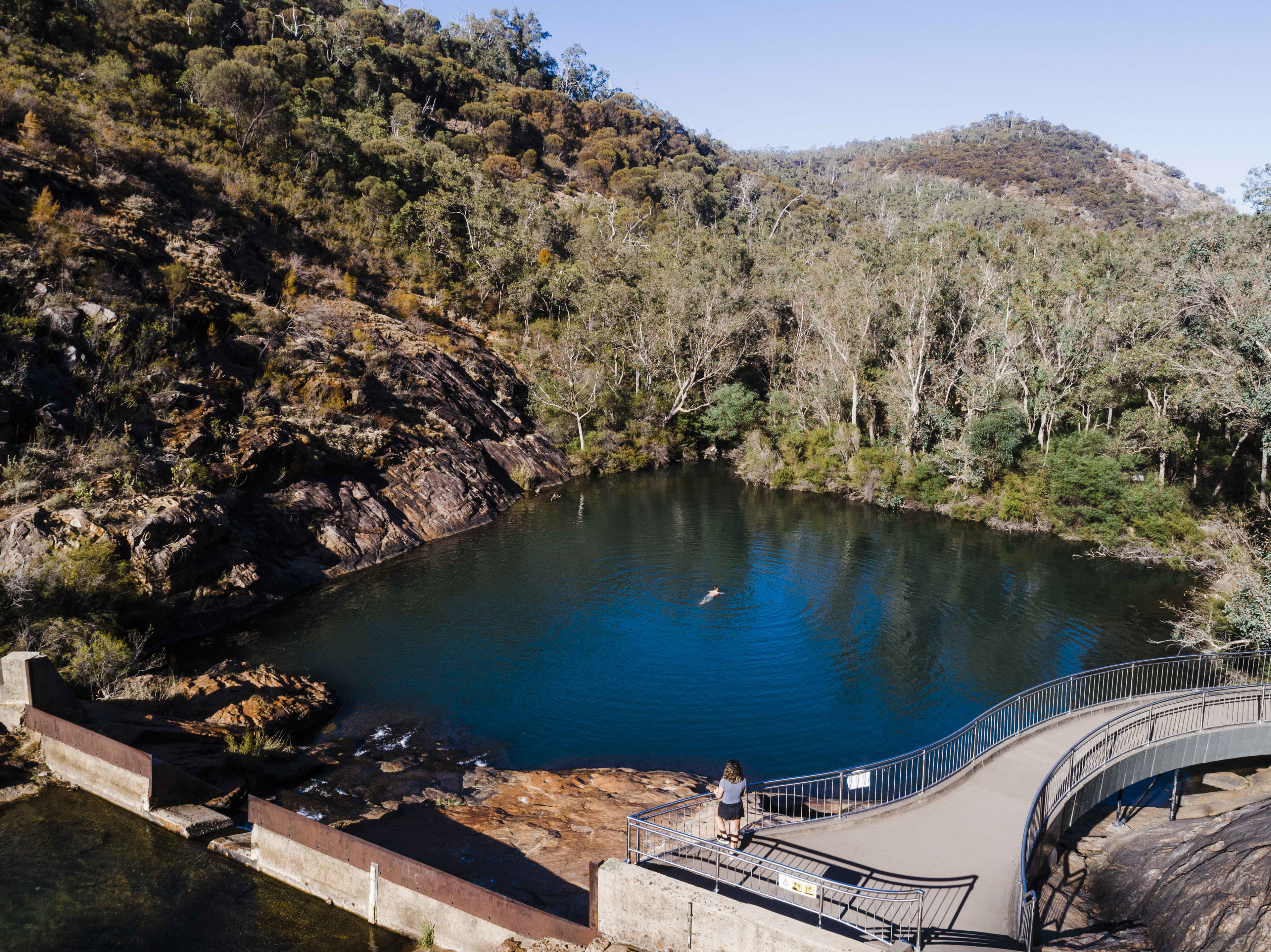 A scenic view of a deep blue natural swimming hole surrounded by rocky cliffs and eucalyptus-covered hills. A lone swimmer is in the center of the water, while a person stands on a curved viewing platform overlooking the pool. Clear blue skies and rugged bushland frame the peaceful landscape.