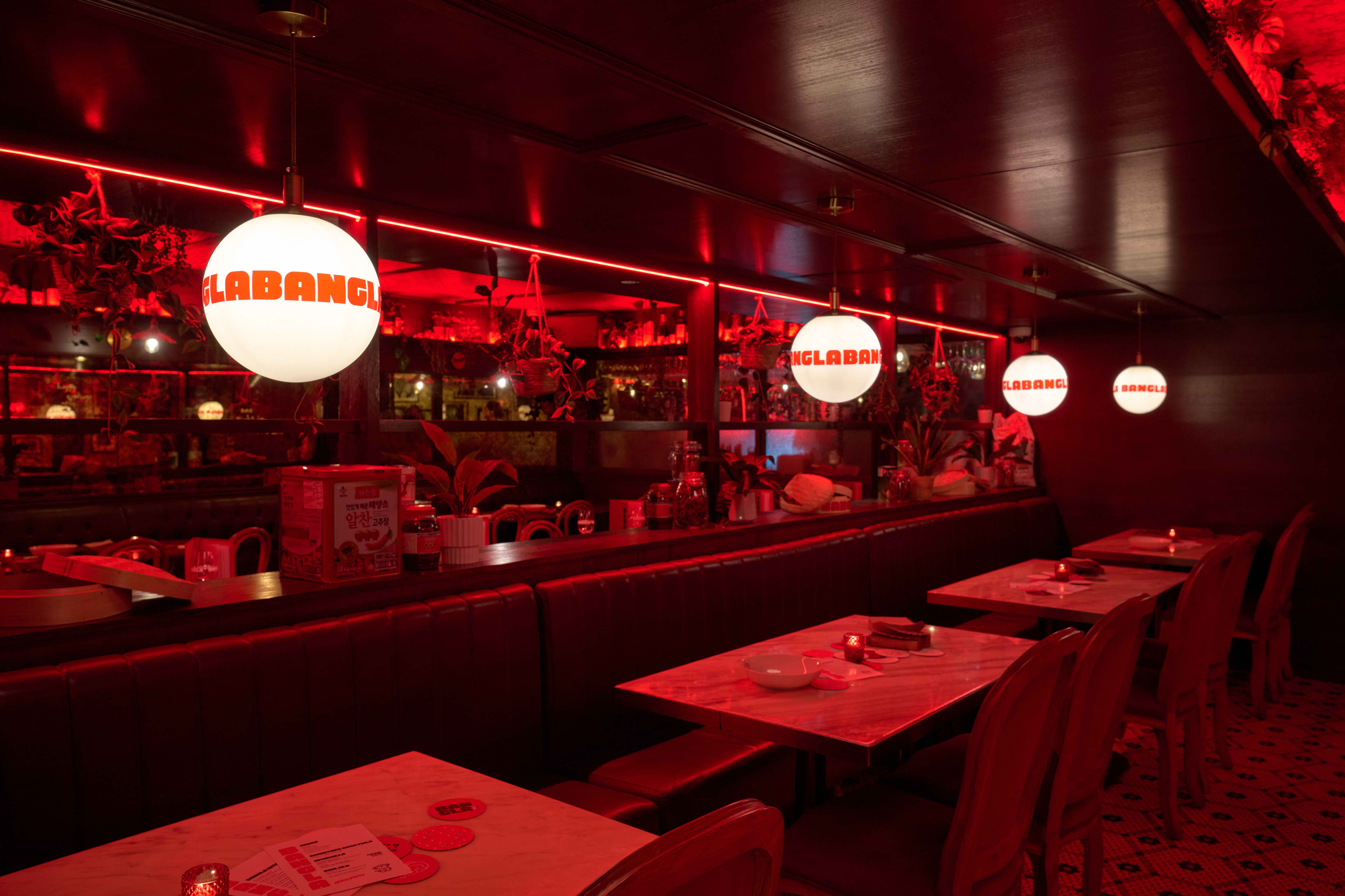 An interior view of a dimly lit restaurant bathed in an intense red neon glow. A row of empty dining tables with marble tops and red chairs lines the right side of the space. Large, spherical white pendant lights hang above the tables, each featuring the bold, black text "LABANGLABANGLABANG" repeated around its circumference.