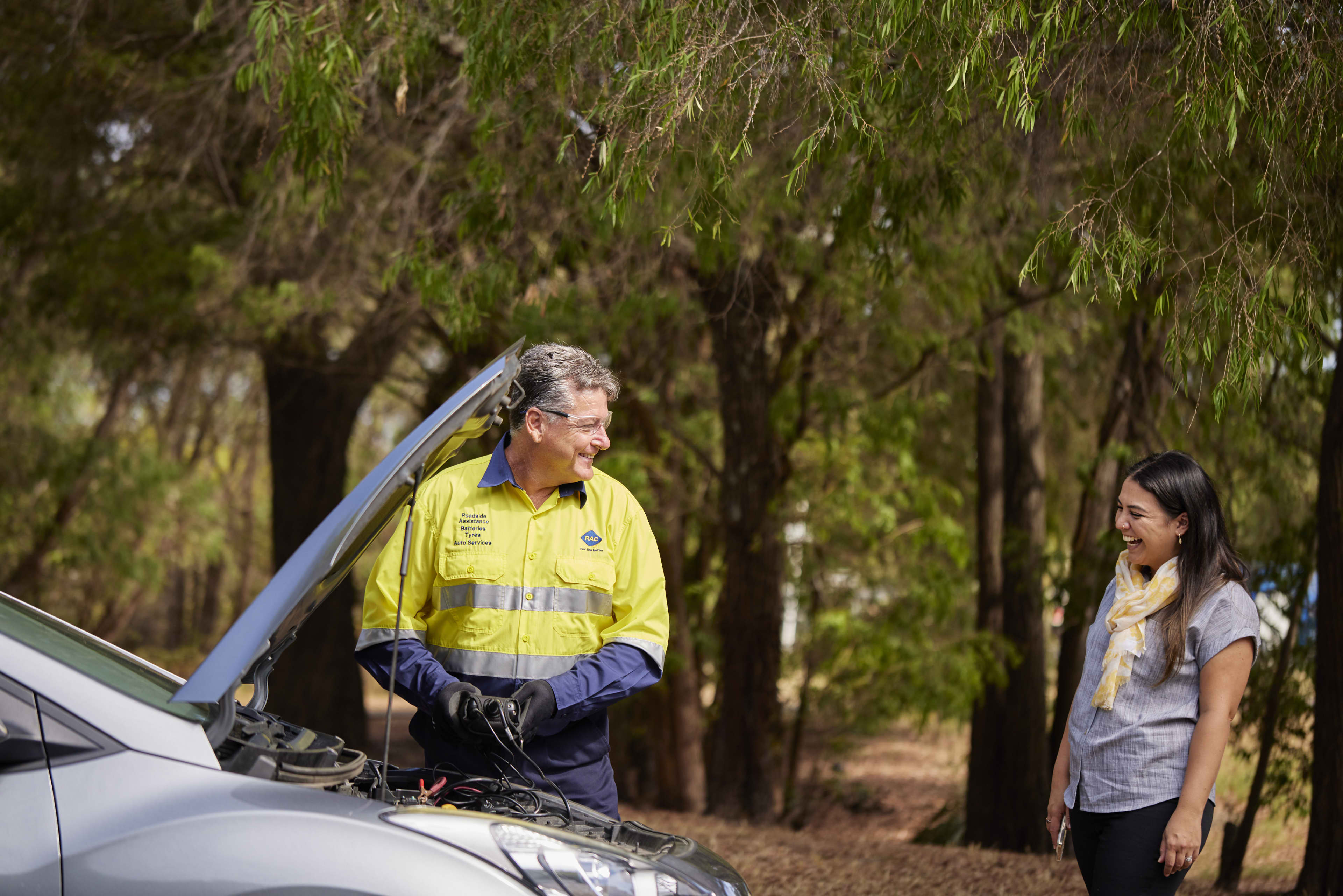 An older male roadside assistance technician wearing a high-visibility yellow and blue uniform, glasses, and black gloves smiles while working under the open hood of a silver car. A young woman with long dark hair, a grey shirt, and a yellow patterned scarf stands nearby, smiling back at him in a wooded, outdoor setting.