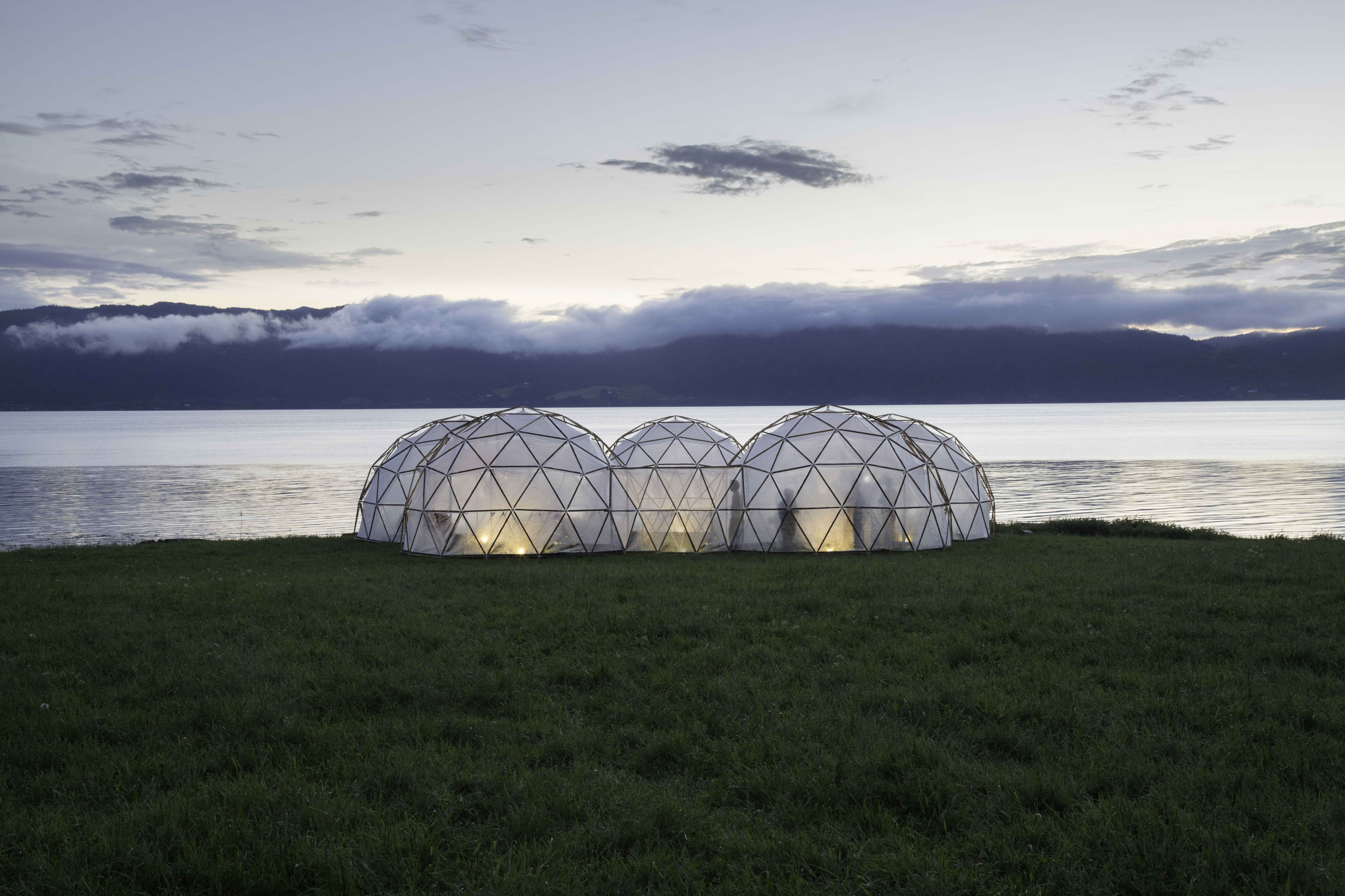 Geodesic dome structures glow at dusk on a grassy lakeshore, with calm water, mist-covered mountains, and a cloudy sky in the background.