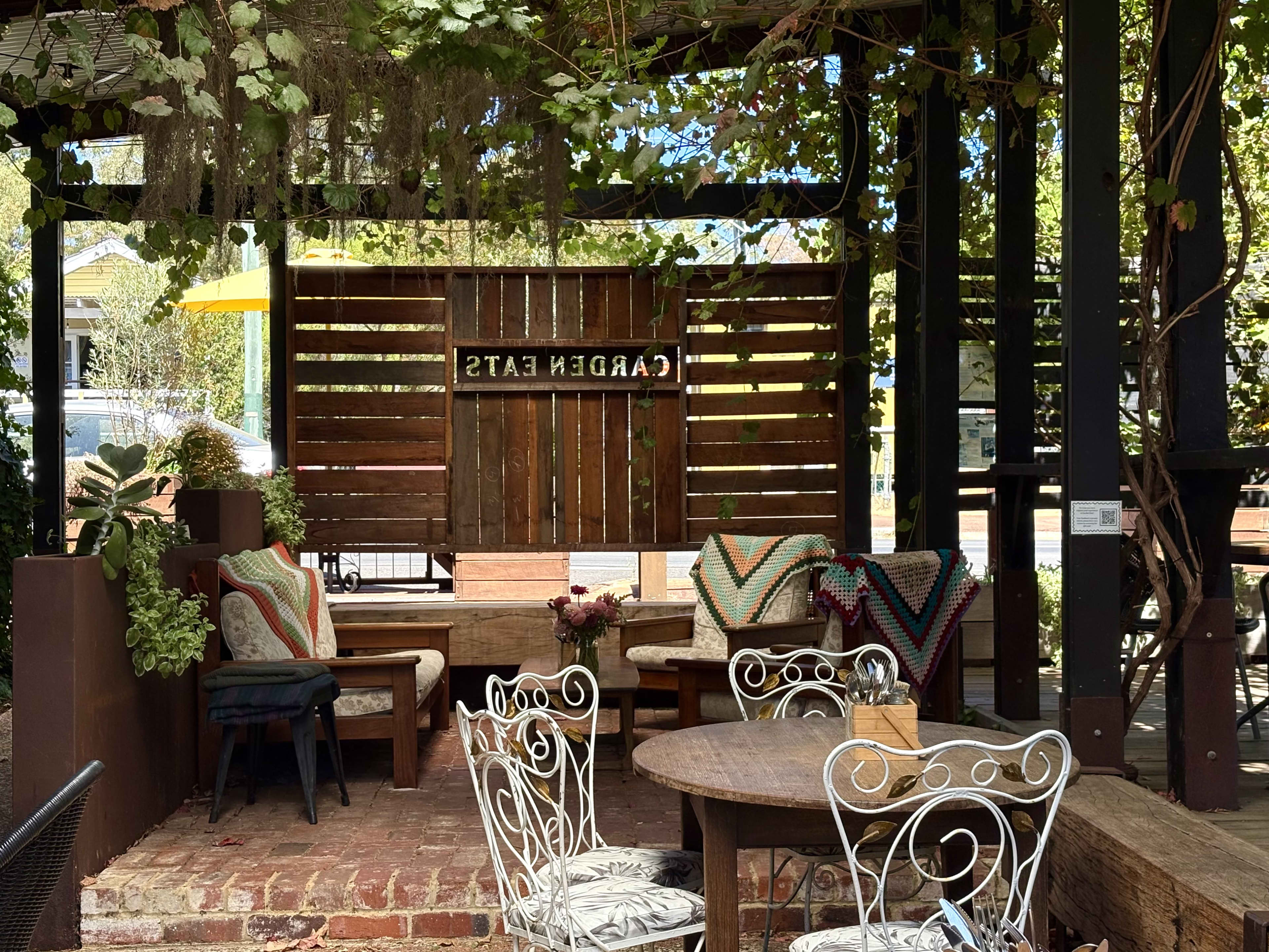 An outdoor café patio with brick flooring featuring a wooden slat wall divider with a backwards sign reading "GARDEN EATS". Lush green vines and spanish moss hang from the overhead wooden pergola. The seating area includes cushioned wooden armchairs decorated with patterned blankets, and a round wooden table surrounded by white wire-framed chairs.