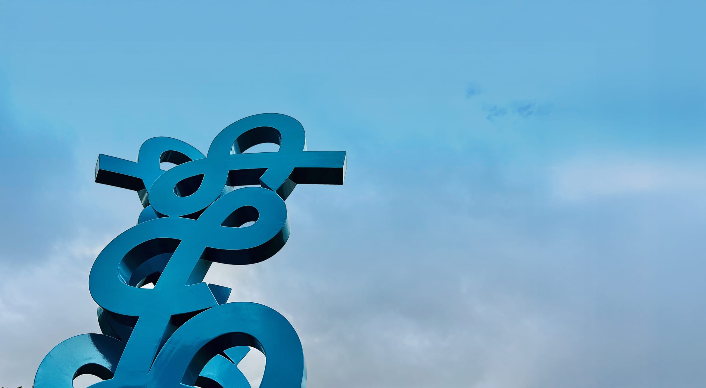 A large, abstract teal-blue metal sculpture made of thick, looping ribbon-like shapes stands against a bright, partially cloudy blue sky. The camera looks upward at the top portion of the modern artwork, highlighting its intertwined geometric curves and sharp metallic angles.