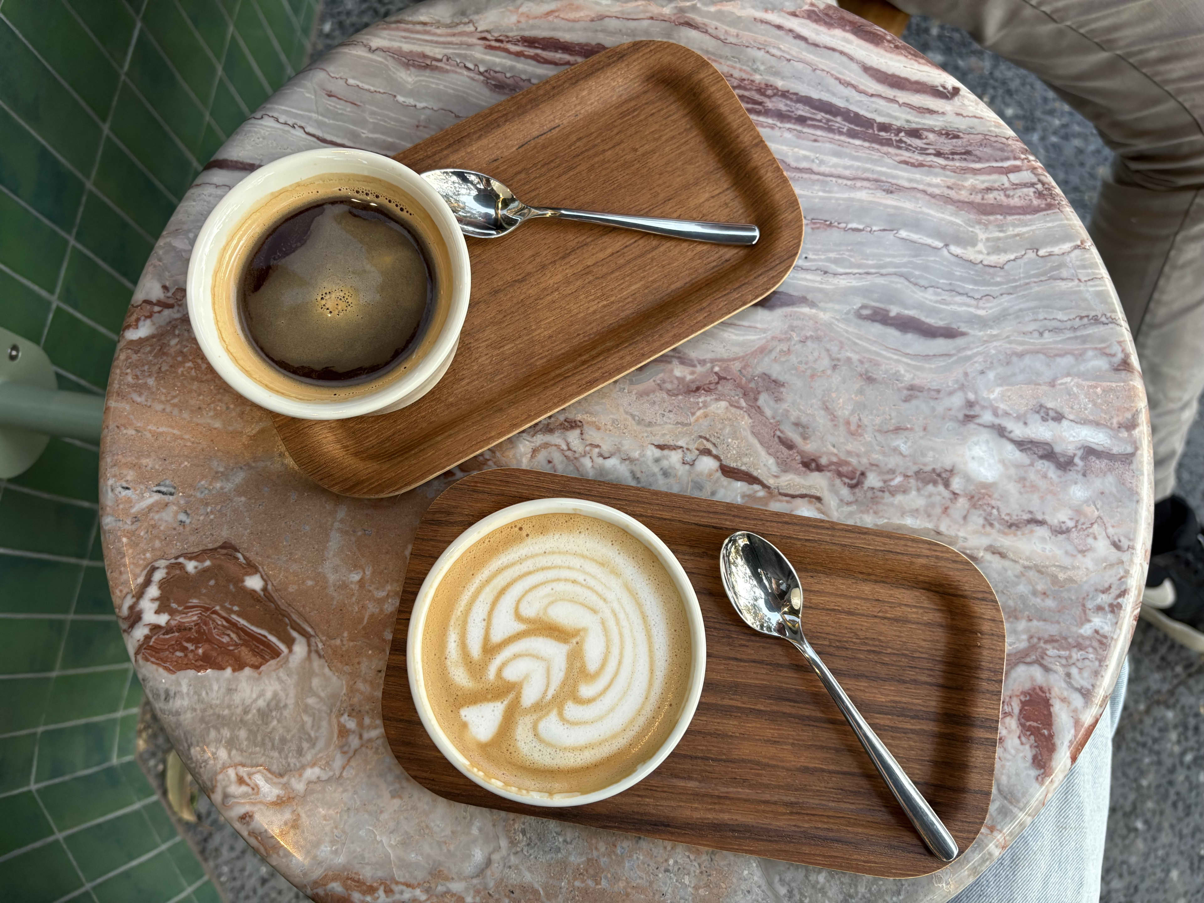 An overhead shot shows two cups of coffee served on individual small wooden trays atop a pink and white marbled table.