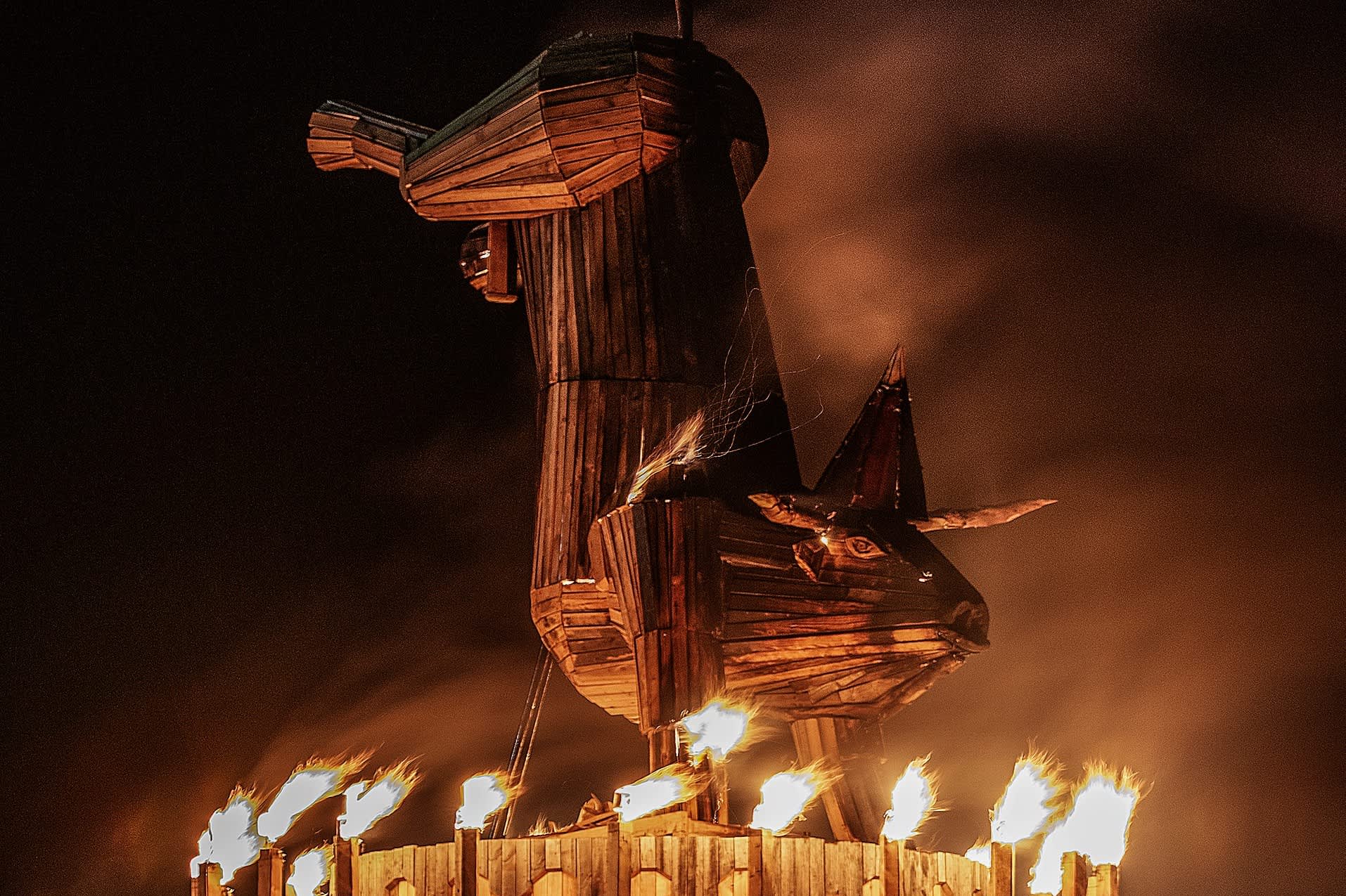 A large, towering wooden sculpture of a bull standing over a fiery base, captured at night.