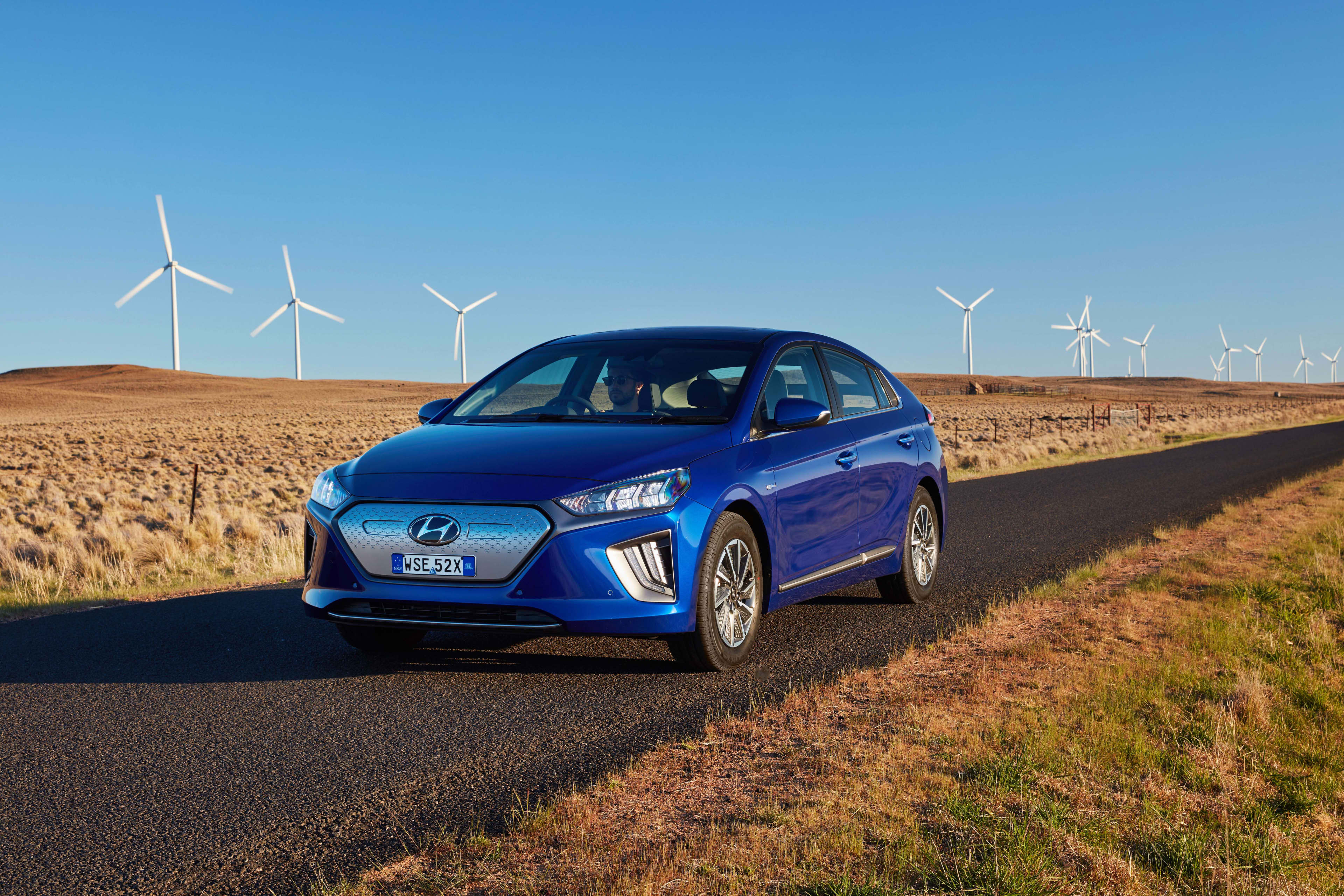 A blue 2019-2022 Hyundai Ionic EV drives on a bitumen road through a landscape of dry grass, with wind power turbines in the background against a blue sky.