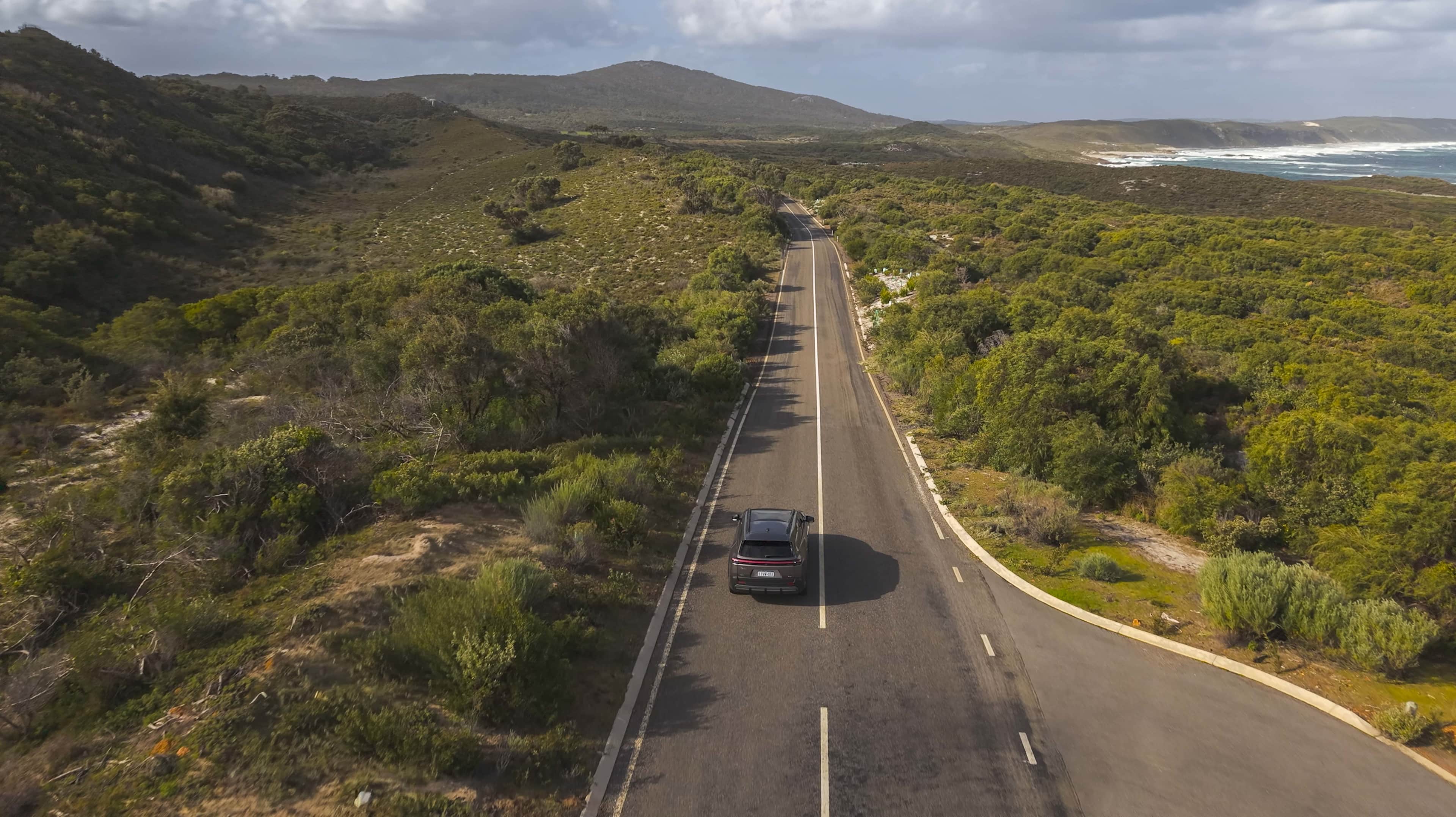 An EV drives down a road in WA's South West, surrounded by greenery and with ocean in the distance.