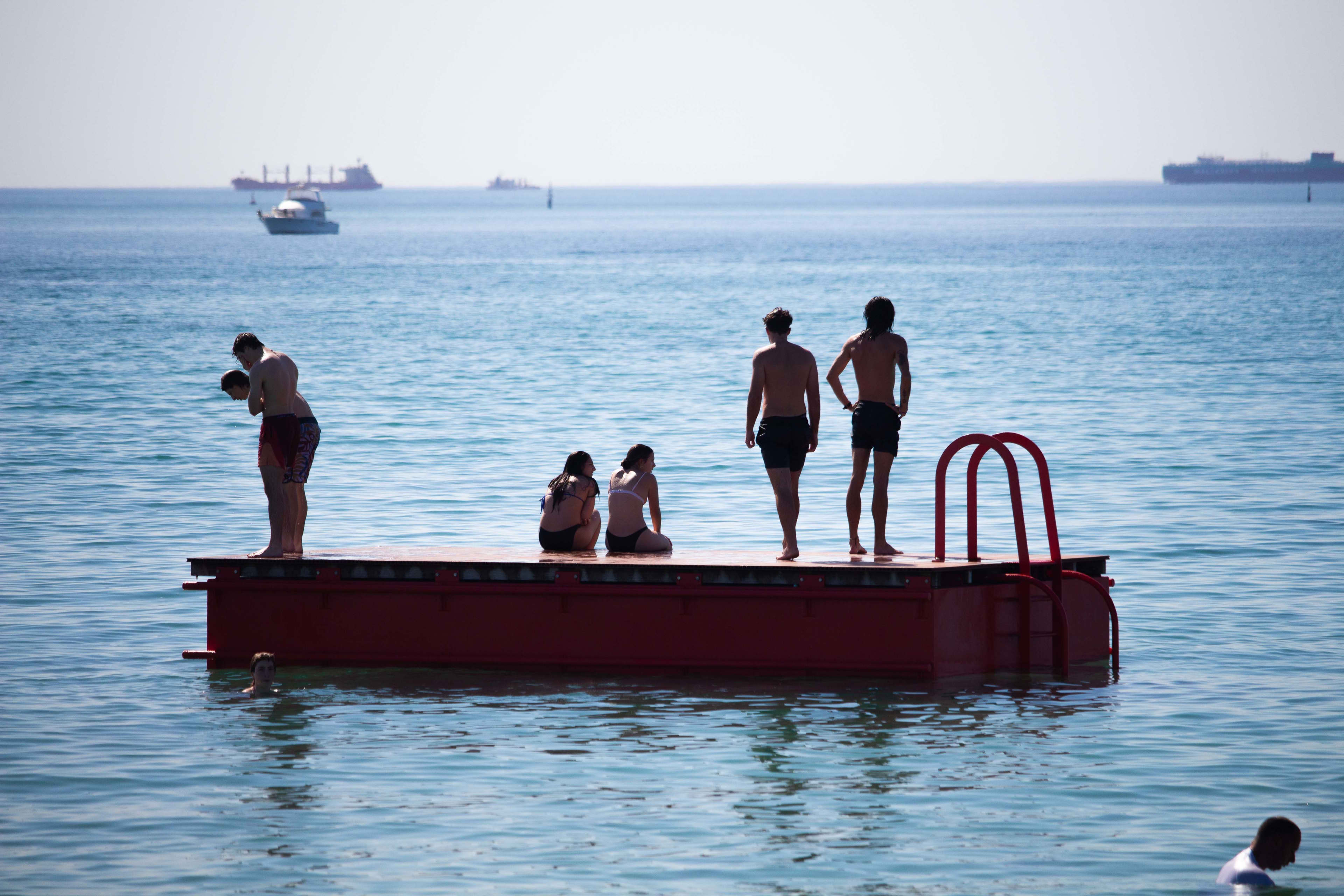 People stand on a red pontoon with a ladder, in a calm ocean with boats in the distance.