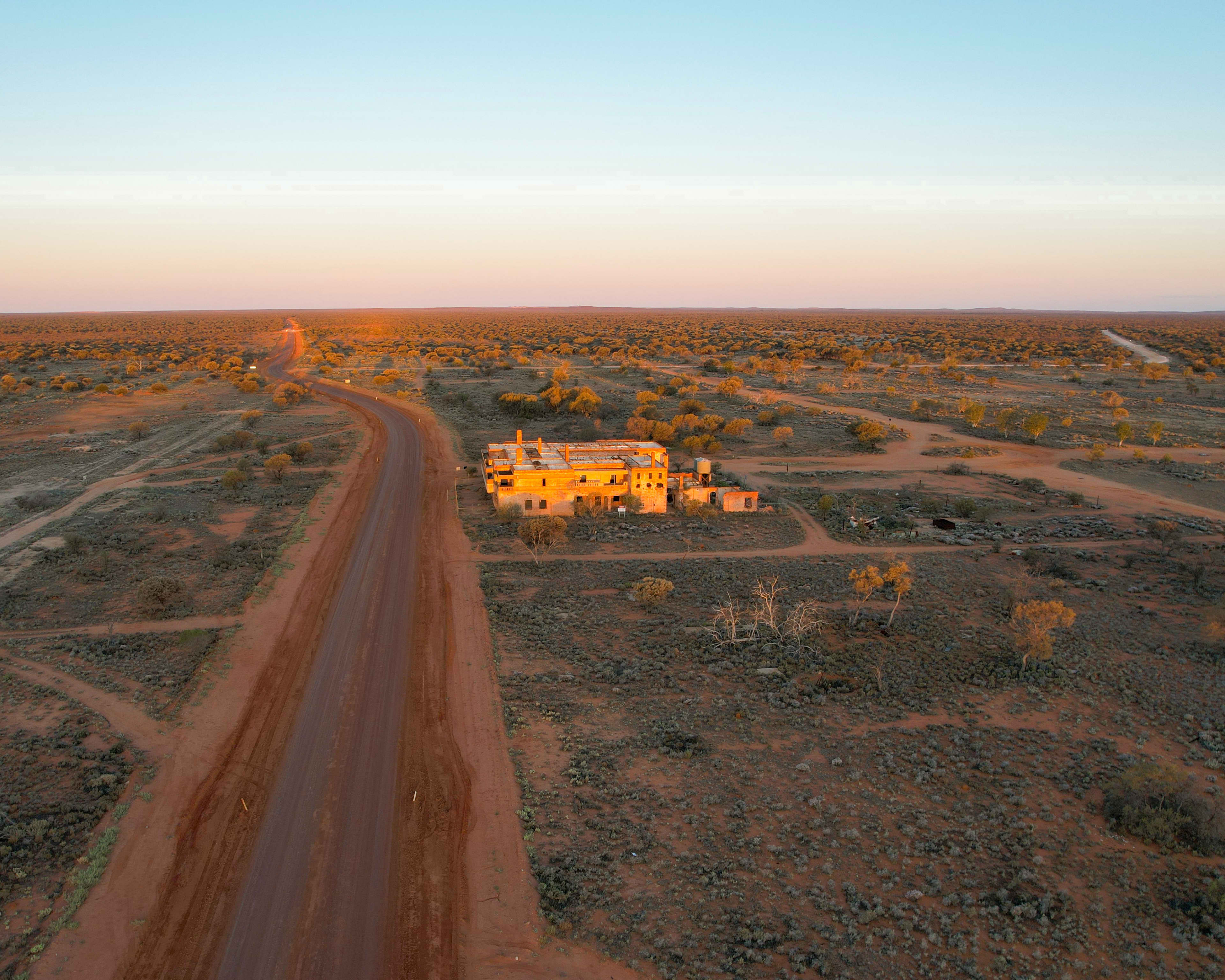 An aerial, wide-angle shot captures a remote building situated in a vast, desert landscape under a clear sky during sunset.