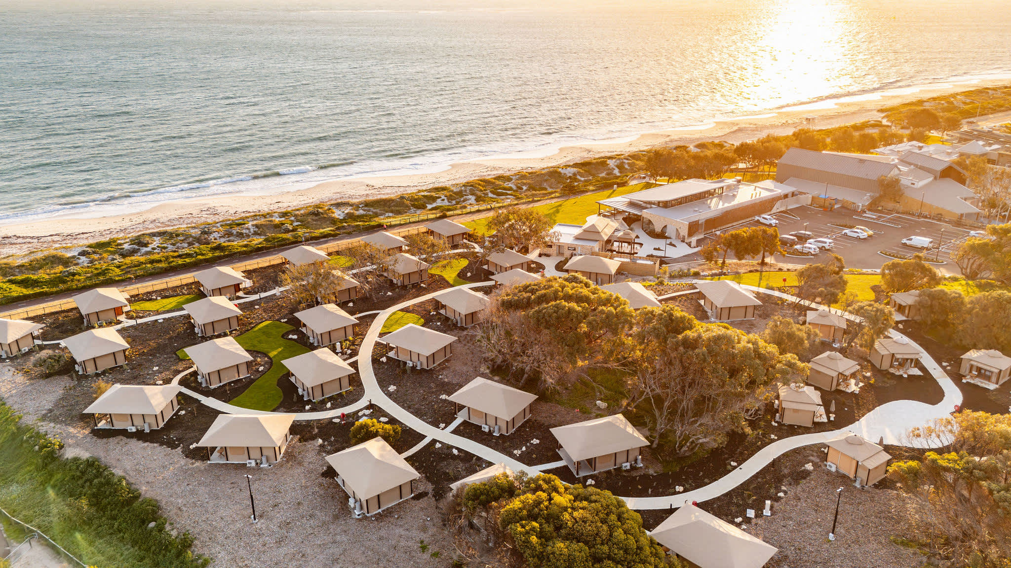 An aerial, high-angle view shows a coastal resort featuring small, detached cabins and chalet nestled on a sandy beach.