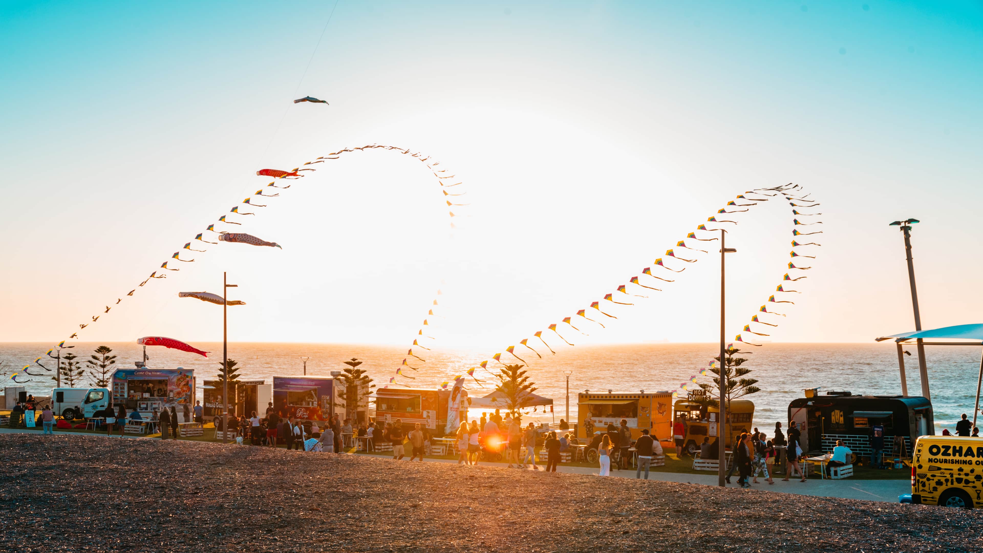 An image at dusk with a line of food trucks sitting in front of the ocean front, with large decorational kites flying in the wind.