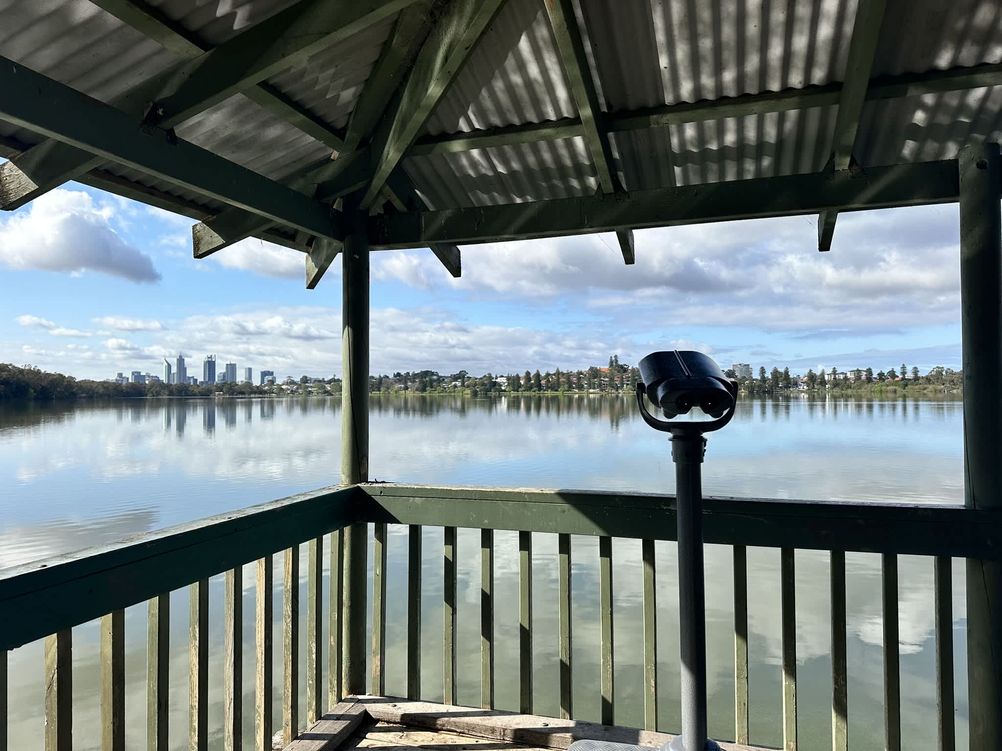 Binoculars inside a gazebo overlooking a lake surrounding by trees with city skyline in background.